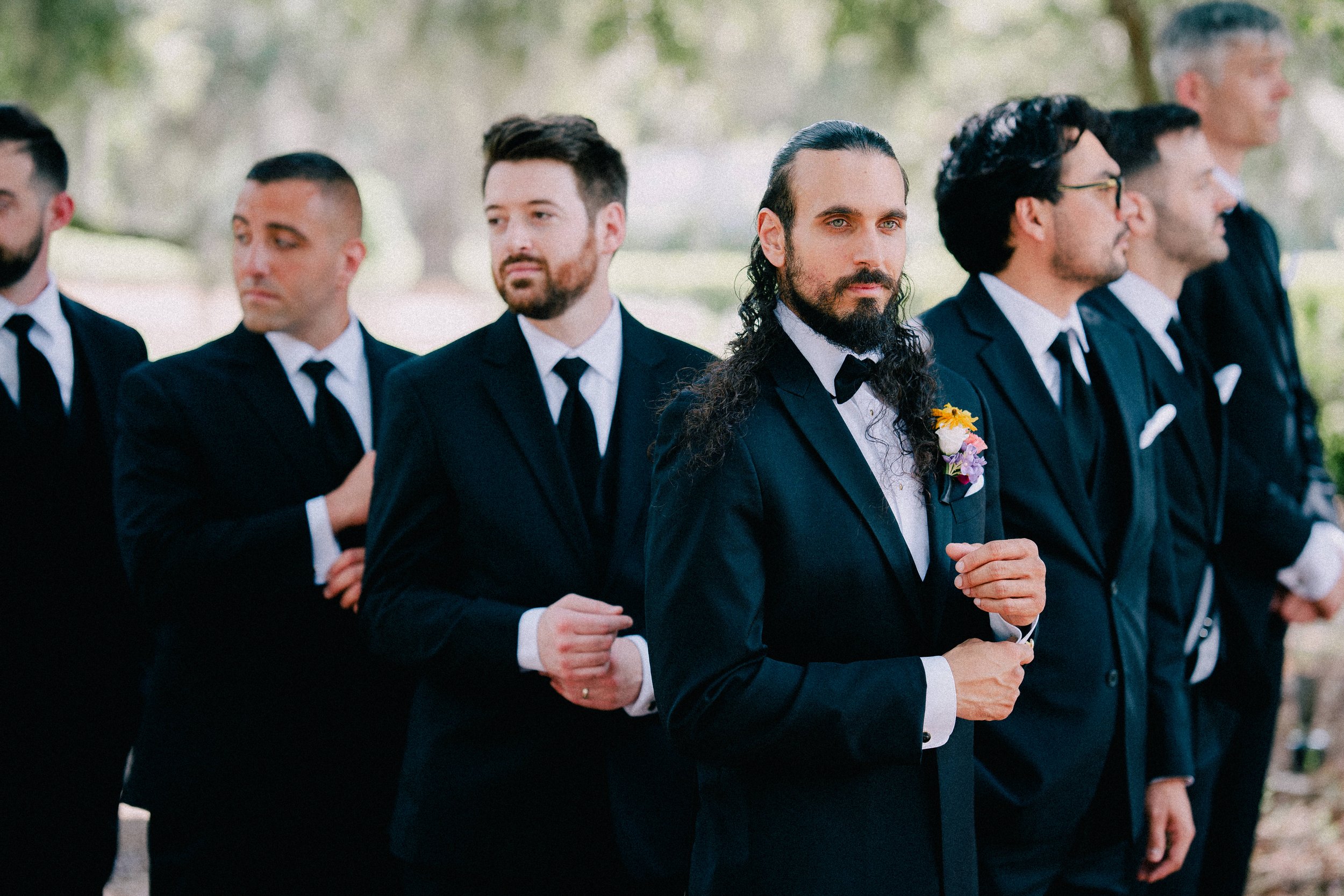 A group of men dressed in formal black tuxedos standing outdoors, likely at a wedding or formal event.