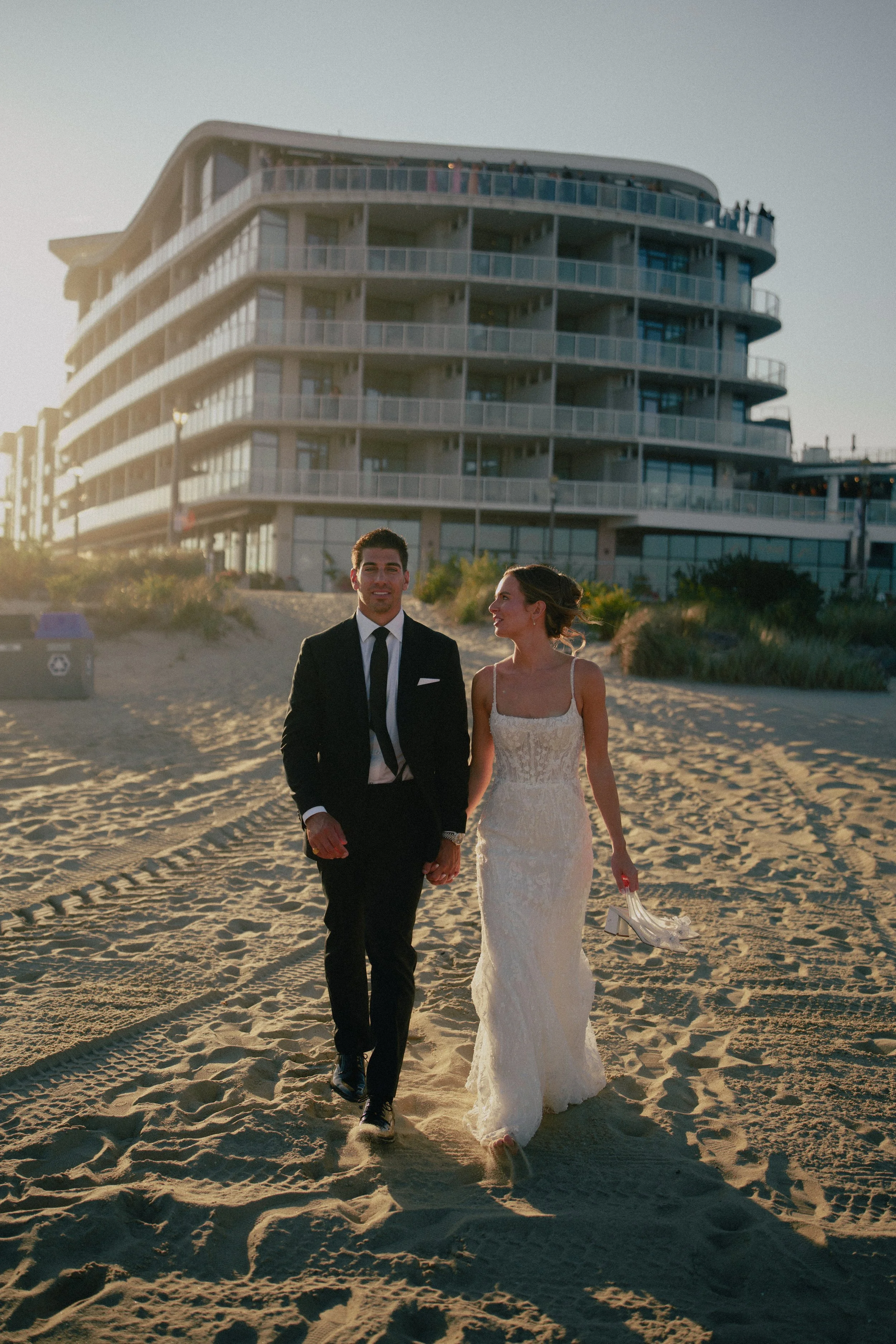 A NJ couple walking on the beach at sunset, with the woman holding her shoes, and a modern, multi-story building in the background at Wave resort - NJ Wedding photographers- Timeline Visuals