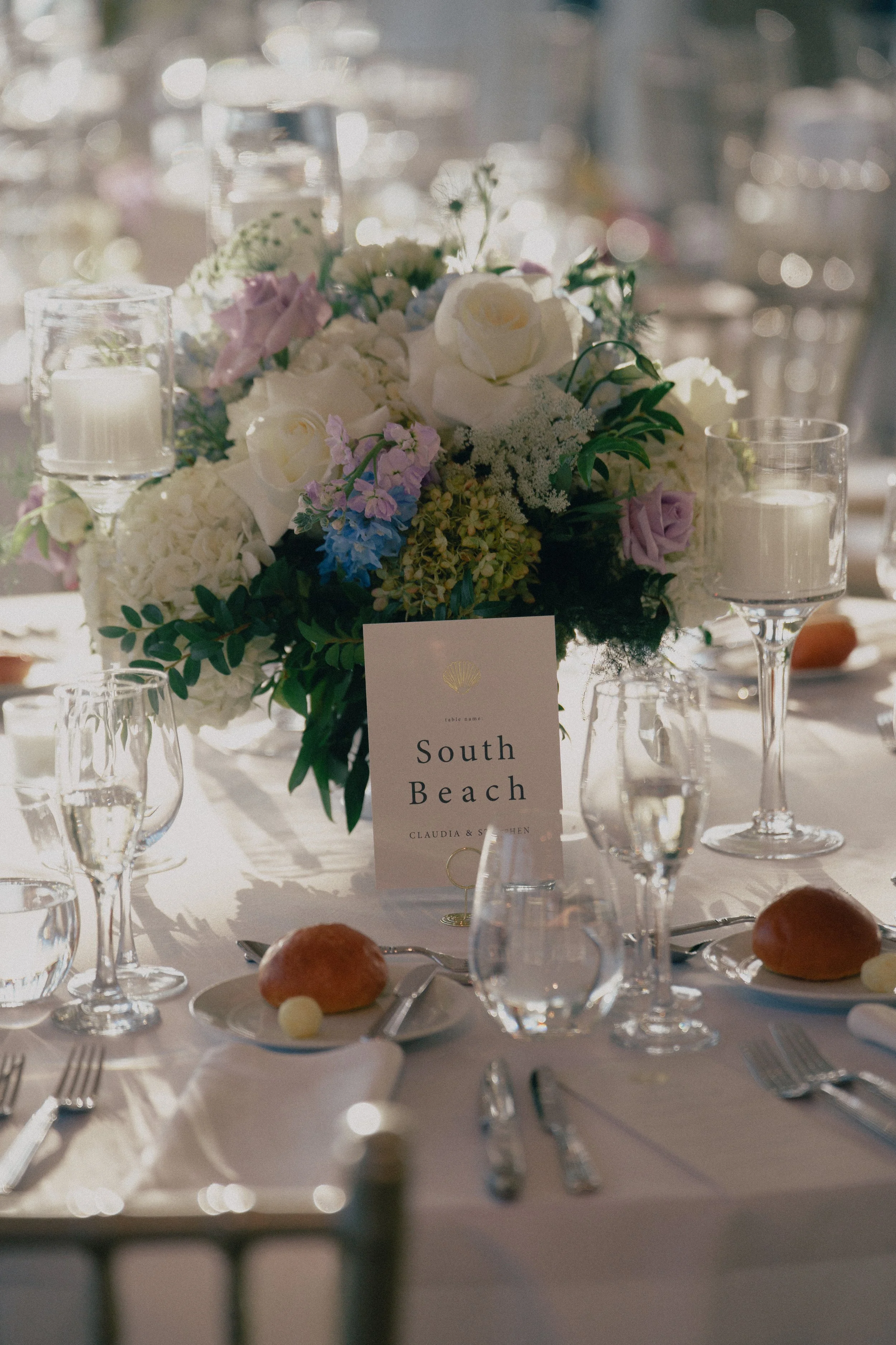 Elegant table setting at a wedding reception with a floral centerpiece, candles, glasses, tableware, and a place card labeled 'South Beach' at Wave resort - NJ Wedding photographers- Timeline Visuals