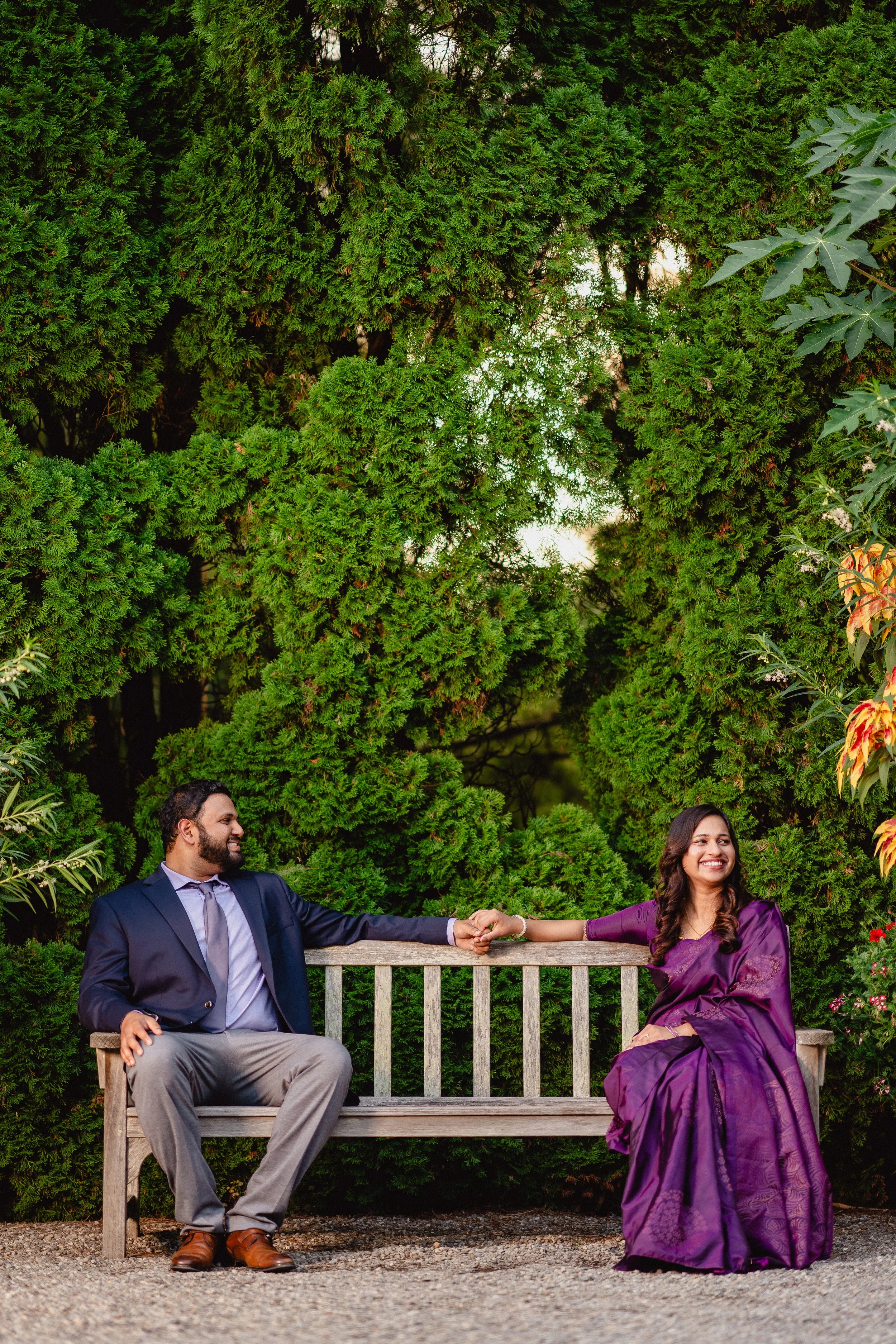 NJ Wedding Photography of A man and a woman are sitting on a wooden bench outdoors, holding hands and smiling at each other, surrounded by lush green trees and plants.