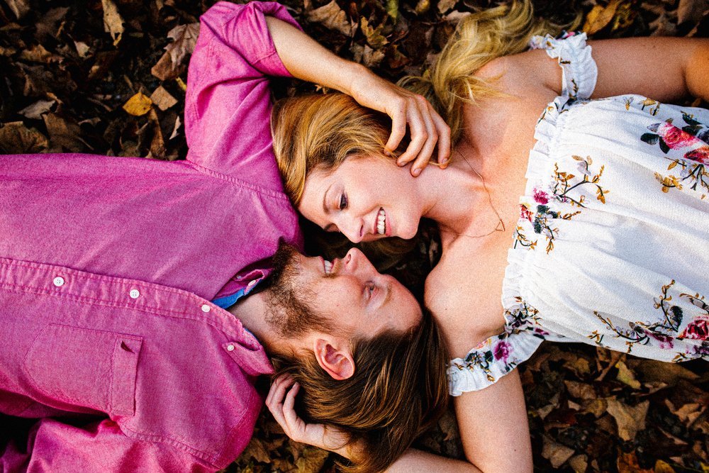 A couple lying on the ground covered with fallen leaves, facing each other and smiling, with the woman's hand gently touching the man's head.