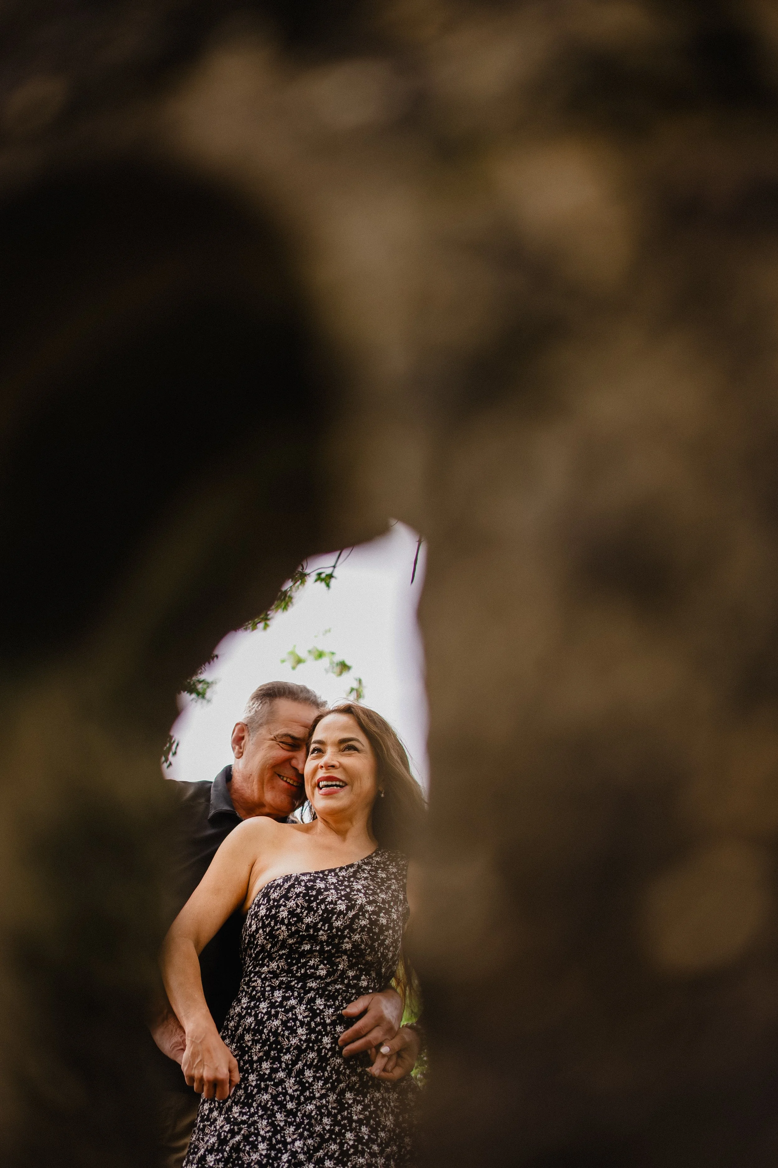 NJ Wedding Photography of A happy couple smiling and embracing each other outdoors, seen through an opening in a tree trunk.