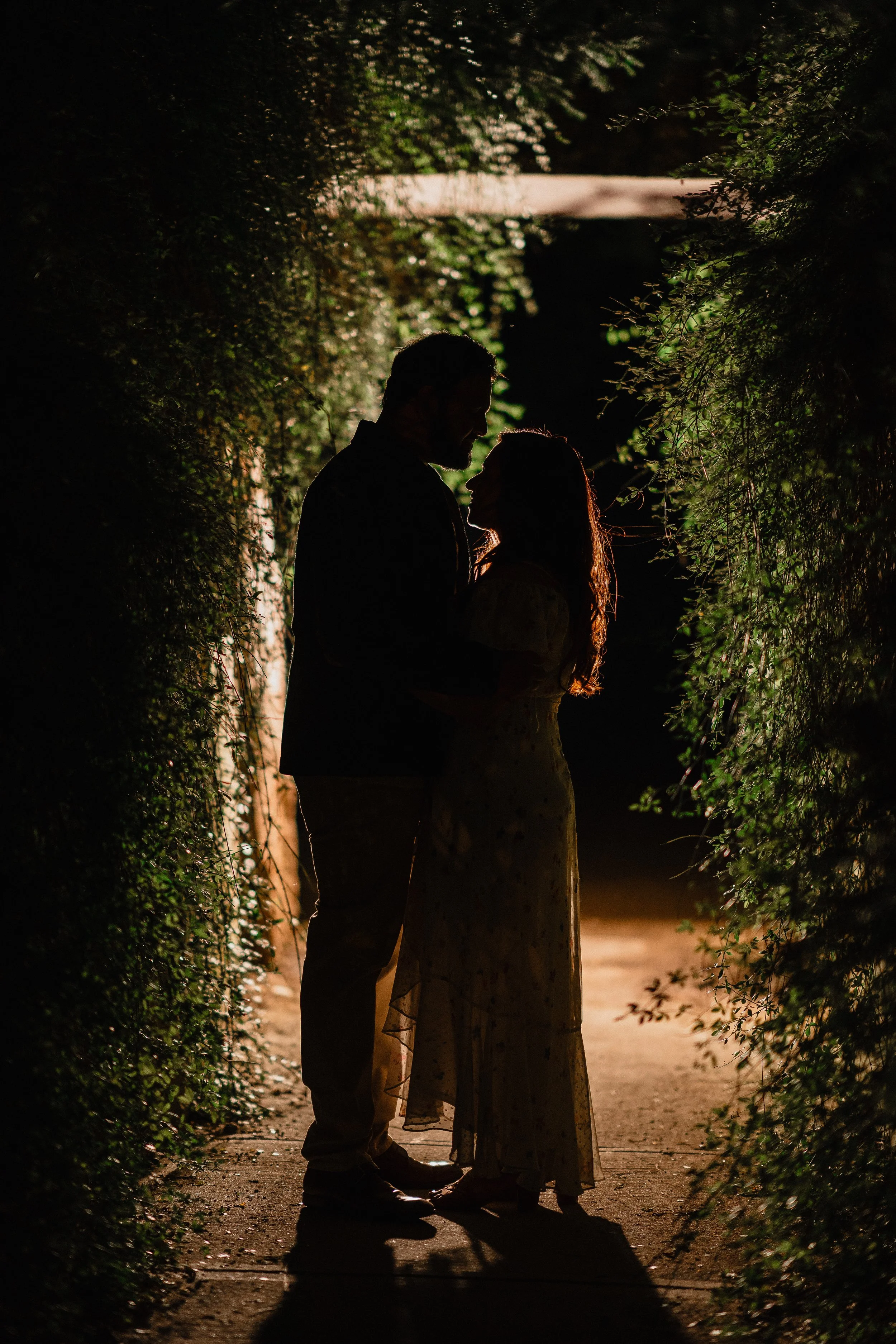 NJ Wedding Photography of Silhouette of a couple standing close together on a dimly lit pathway surrounded by lush greenery, with a light shining from behind them.