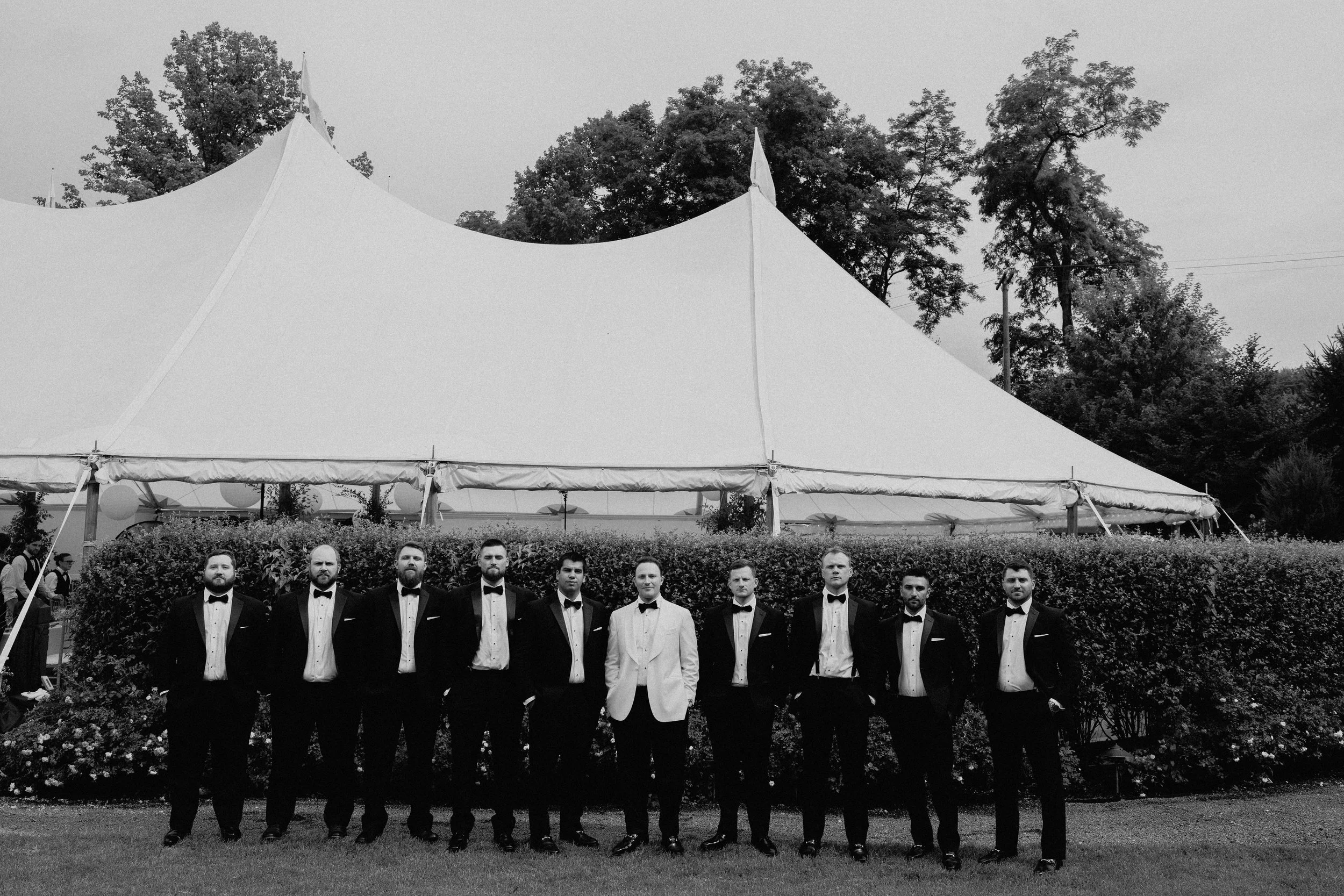 Editorial wedding photography portrait of groomsmen in black tuxedos beside a hedge with the white reception tent behind them.
