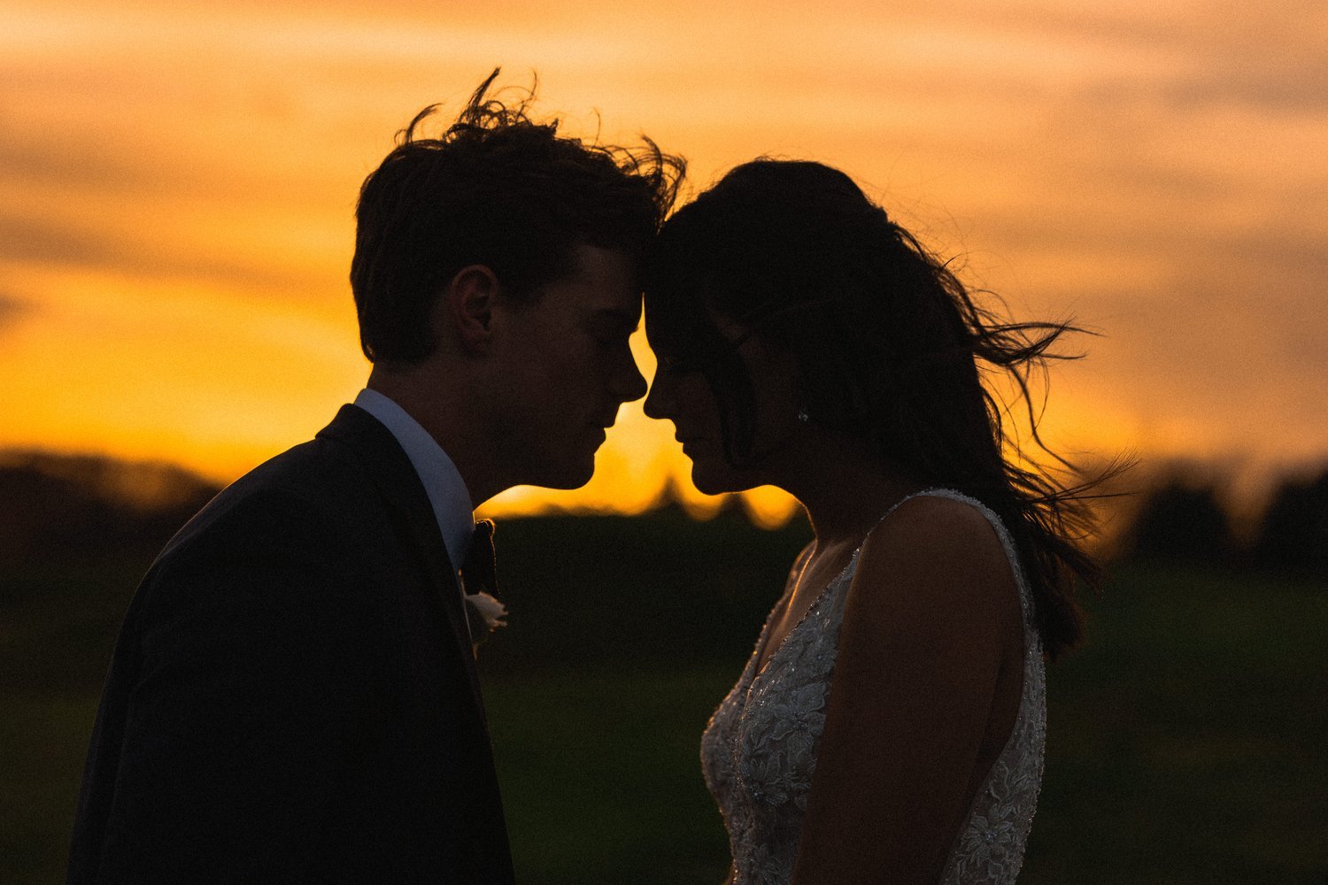 Silhouette of a couple, a man in a suit and a woman in a wedding dress, touching foreheads against a sunset background.