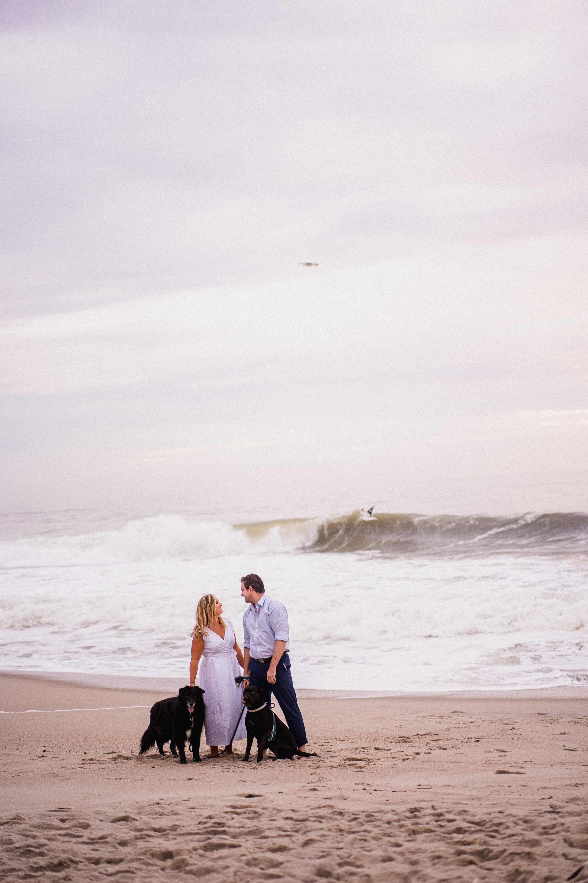 A couple and their two dogs standing on the beach, with waves and surfers in the background, during an overcast day.