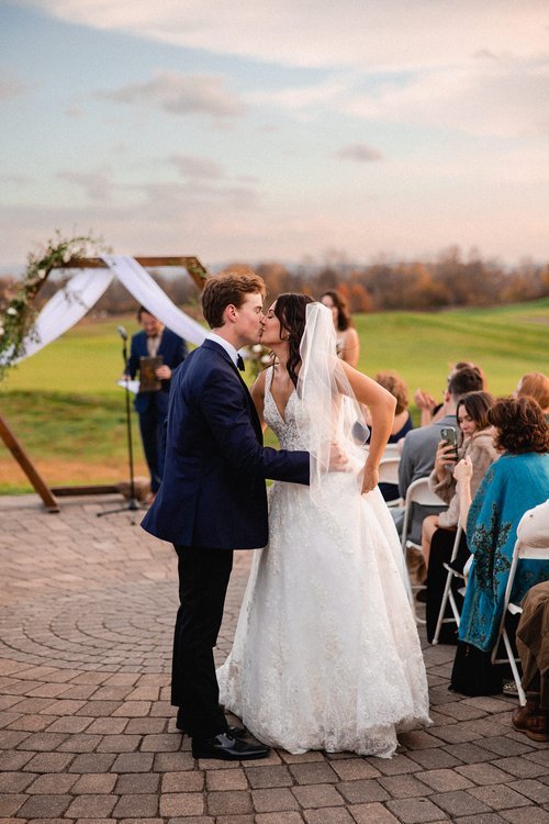 A newlywed couple shares a kiss during their outdoor wedding ceremony, with guests watching and taking photos behind them, under a decorated arch on a brick patio with a scenic green field and cloudy sky in the background.
