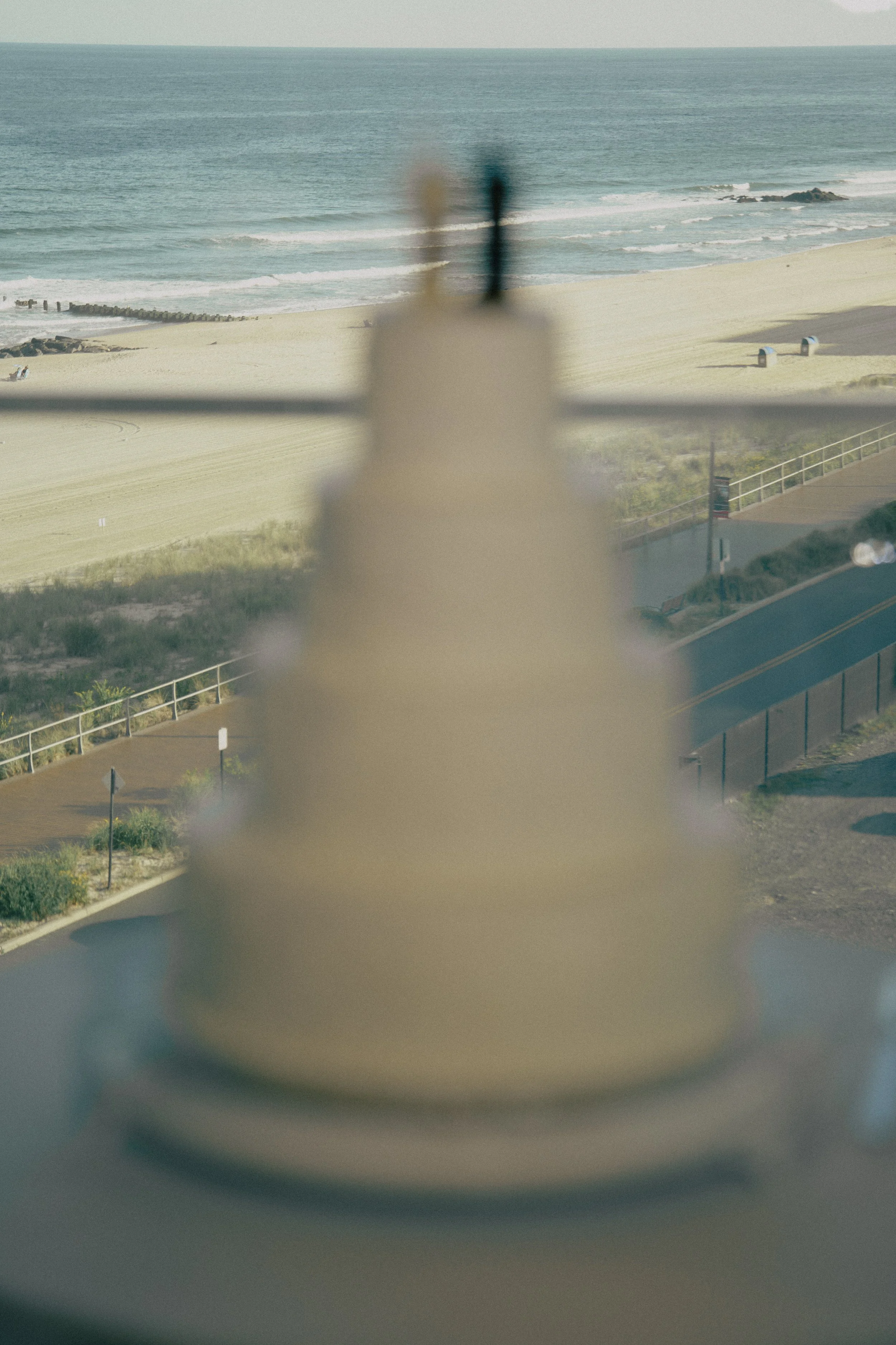 Past a wedding cake blurry view through a window of a sandy beach with a walkway, ocean, and sky in the background at Wave resort - NJ Wedding photographers- Timeline Visuals