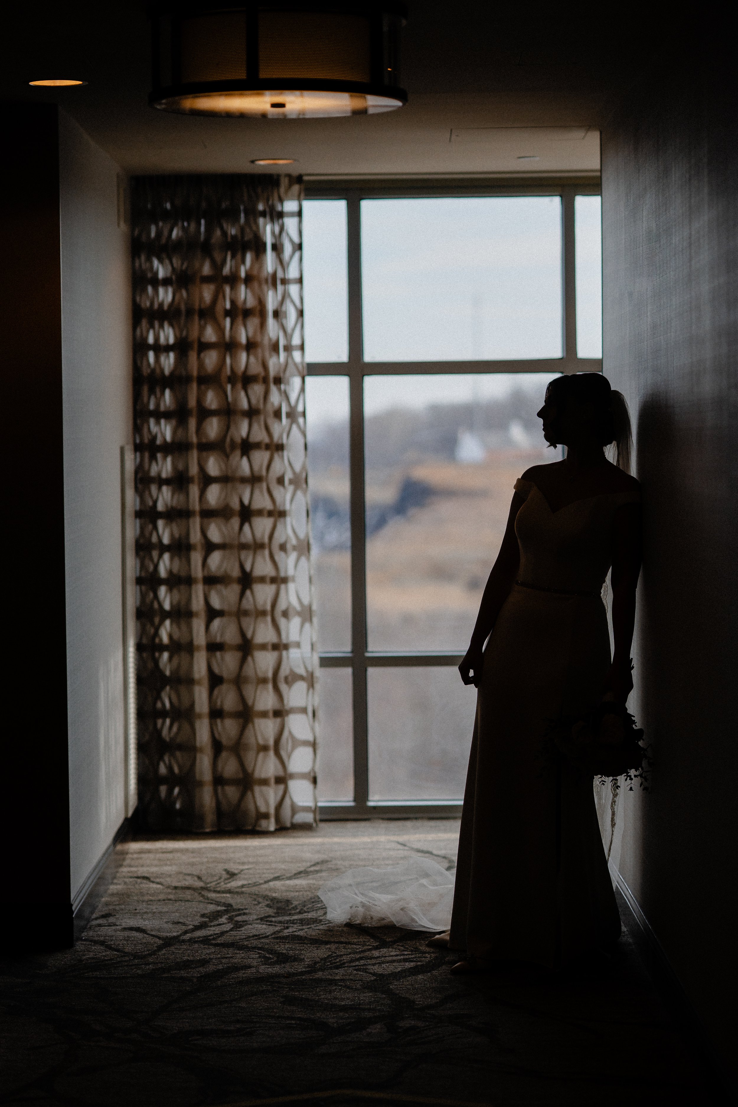 Silhouette of a woman in a wedding dress holding a bouquet, standing against a window with patterned curtains, in a dimly lit room.