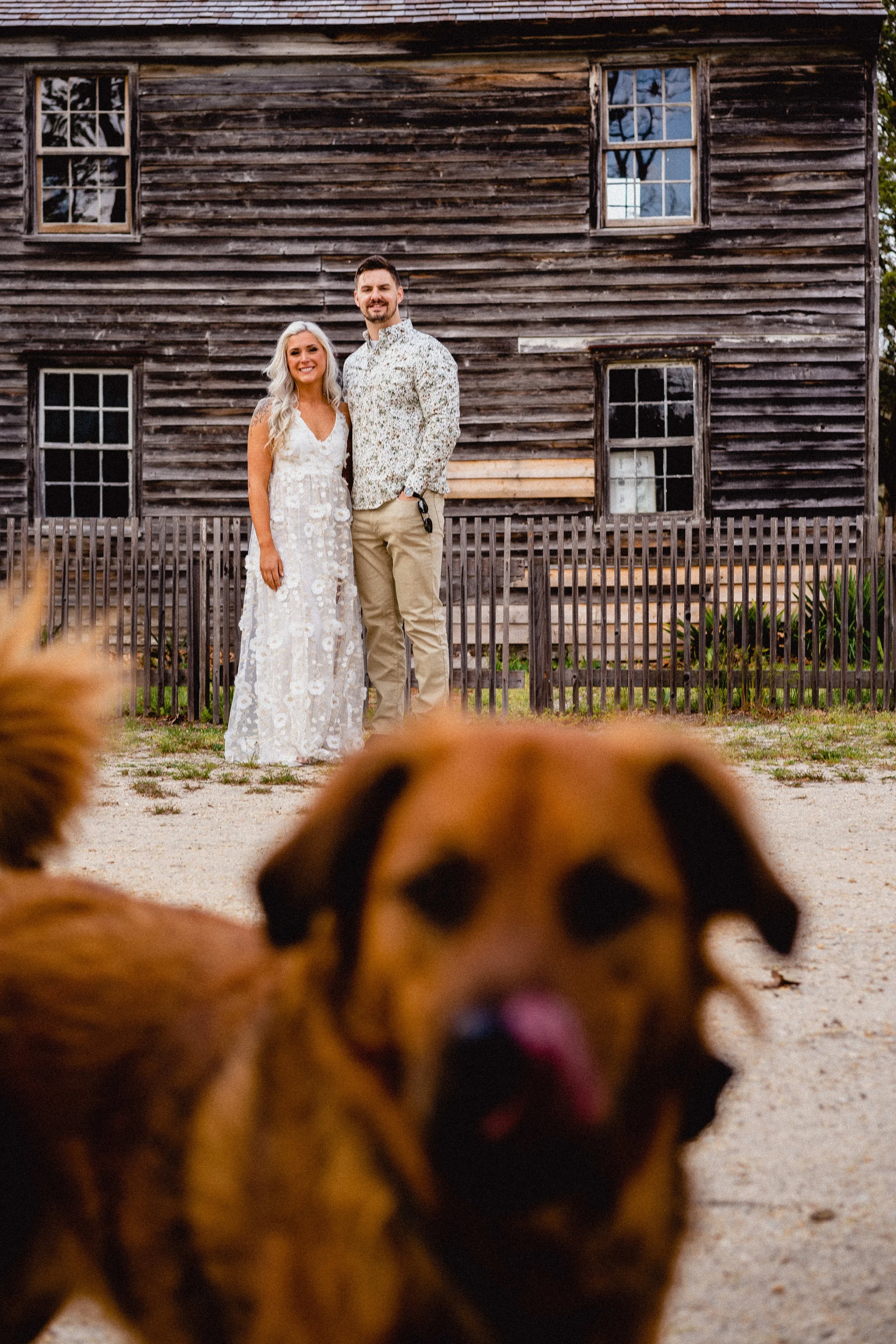NJ Wedding Photography of A couple standing outdoors in front of a rustic wooden building, with a large brown dog in the foreground.