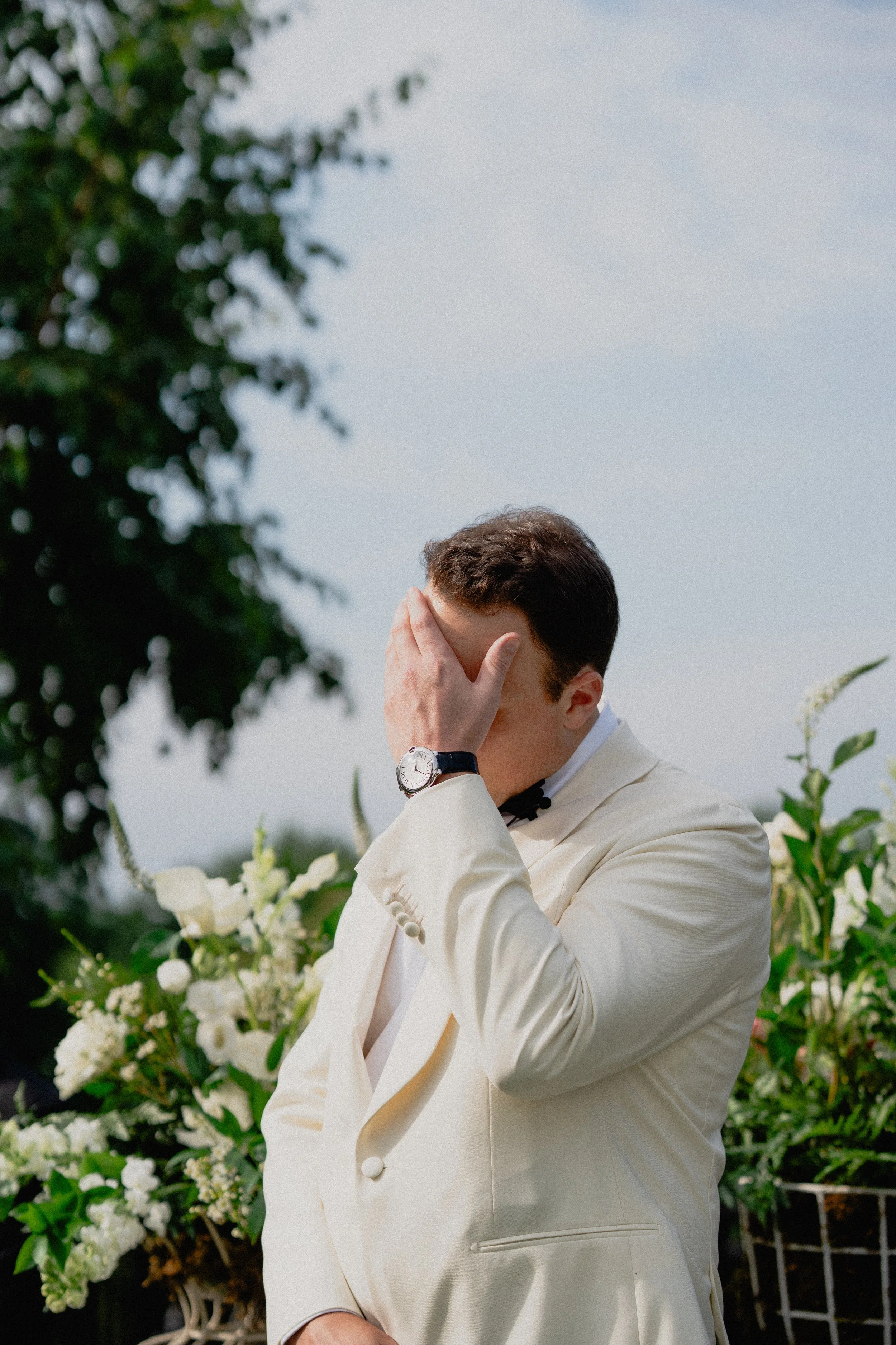Documentary wedding photography captures a groom in a white dinner jacket covering his face with emotion beside ceremony flowers.