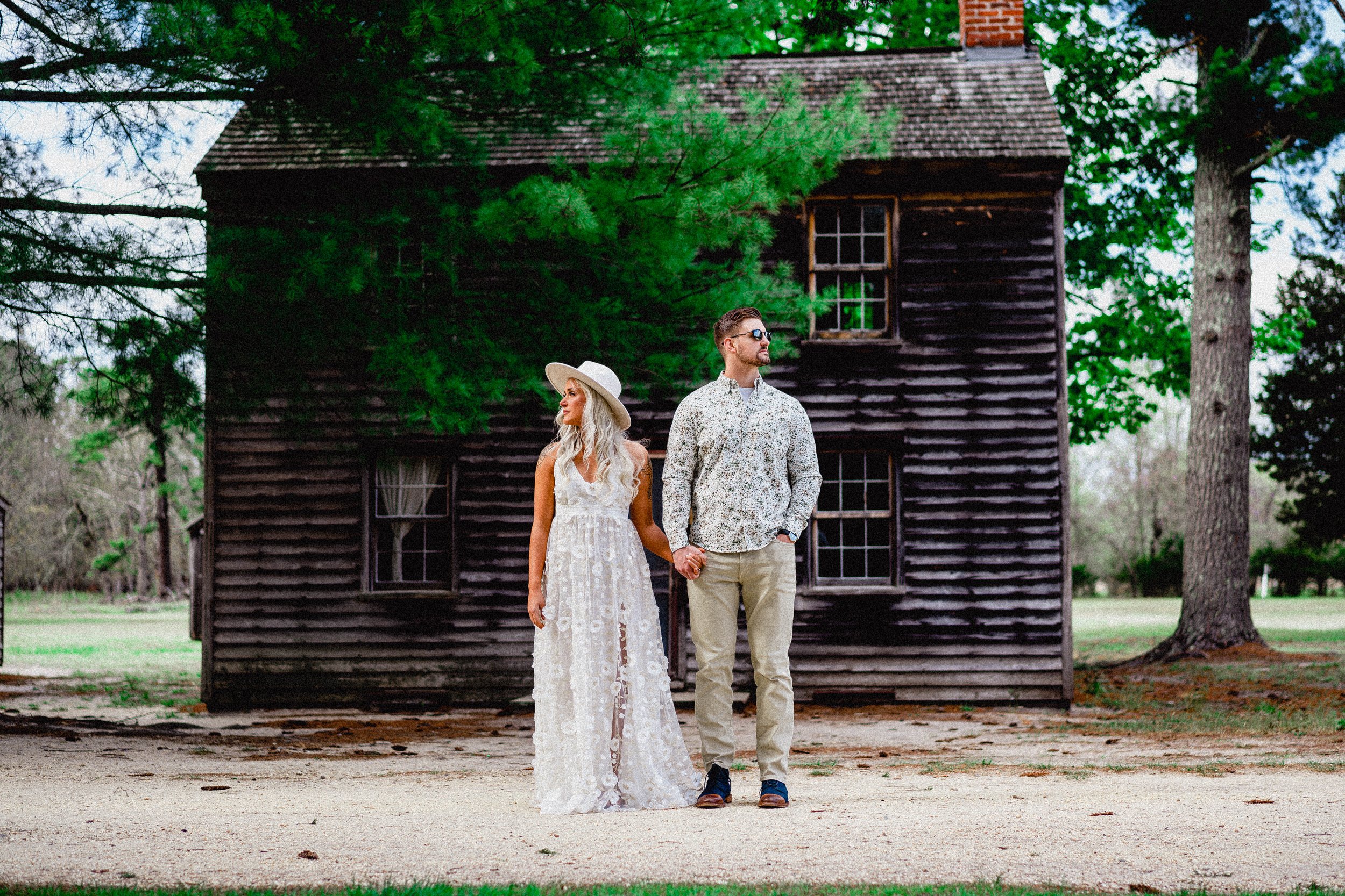 NJ Wedding Photography of A couple holding hands, standing outdoors in front of an old black wooden house with trees around, during daytime.