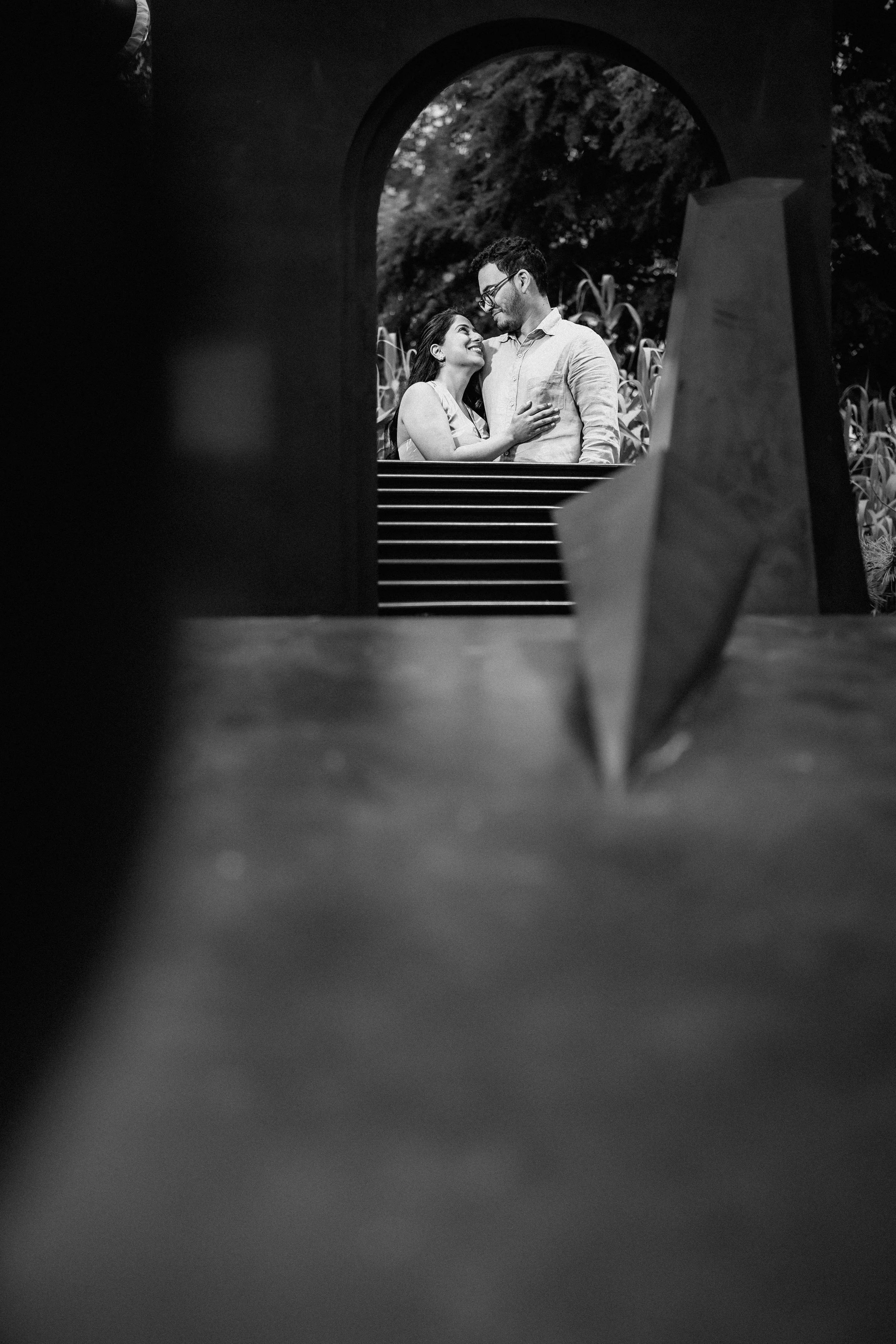 NJ Wedding Photography of A couple is seen through a rectangular opening, smiling and looking at each other, with a staircase and trees in the background.