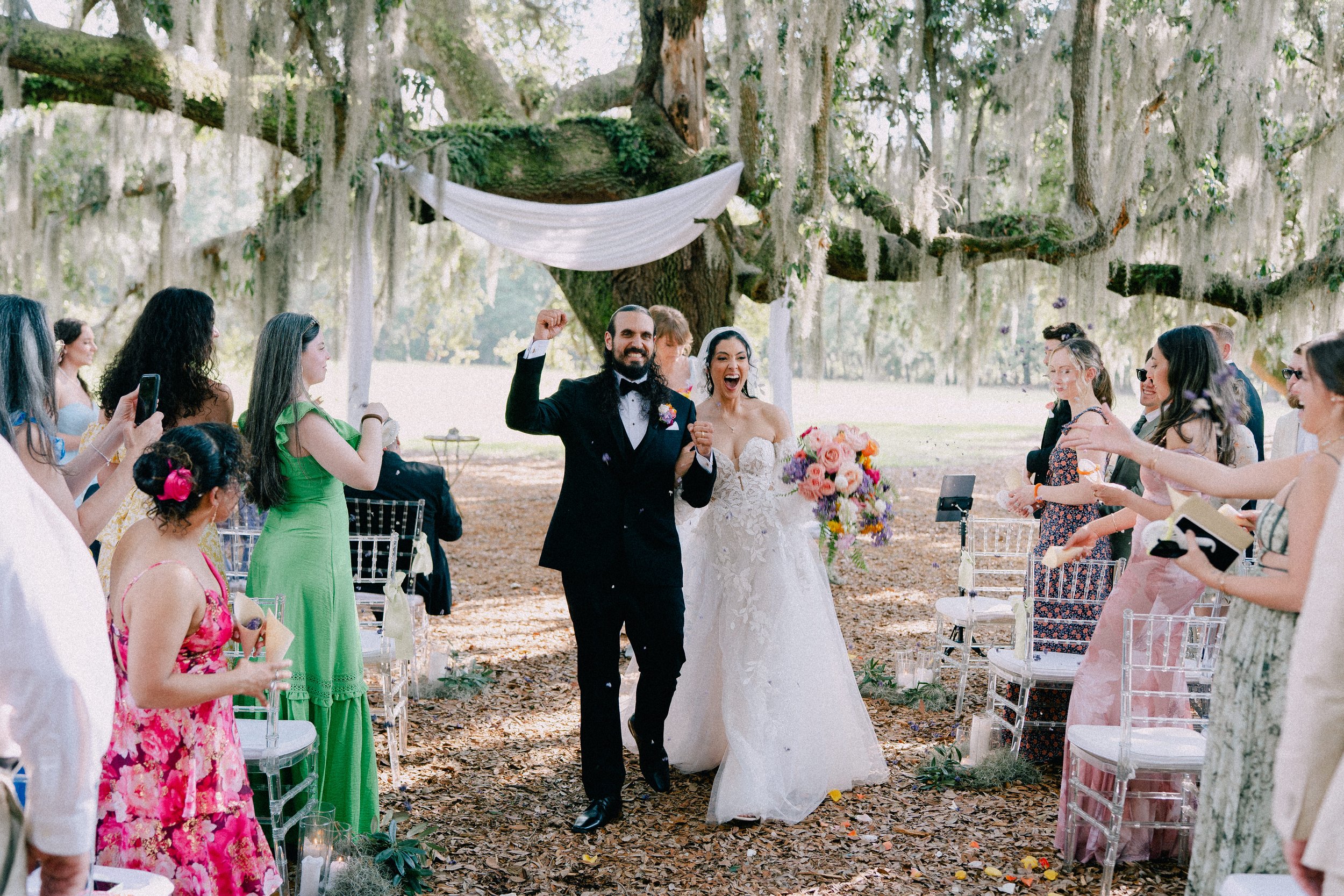 A bride and groom celebrate as they walk down the aisle after their wedding ceremony outdoors surrounded by friends and family in a wooded area with Spanish moss.