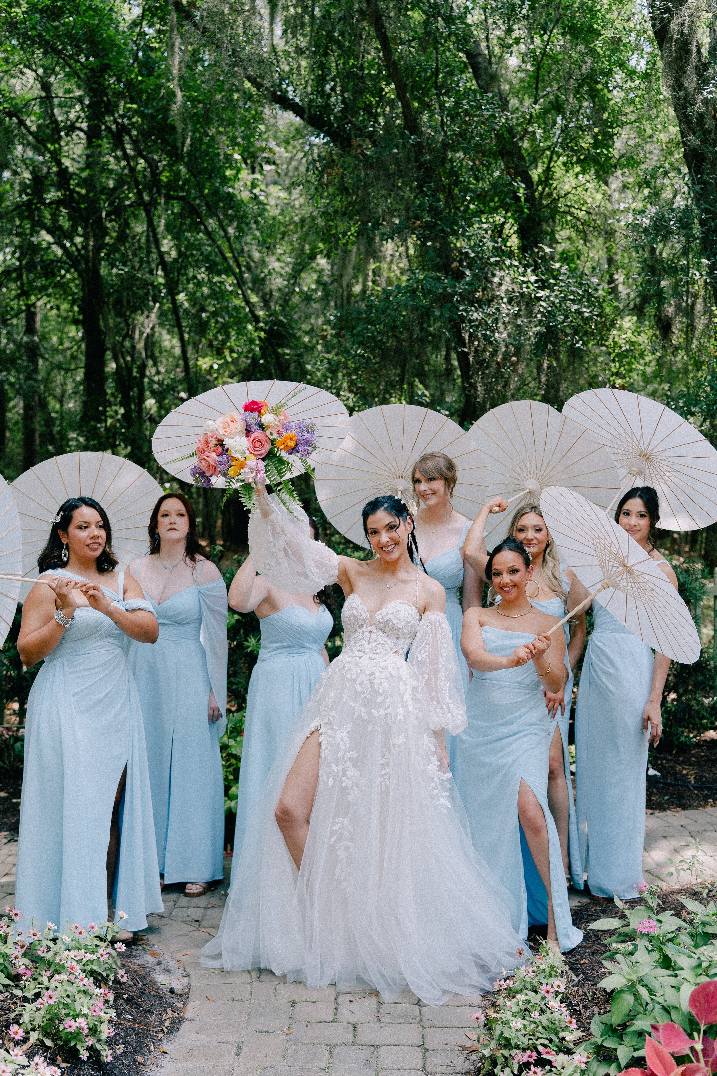 A bride in a white wedding dress holding a bouquet, standing among bridesmaids in light blue dresses, each holding white parasols with a lush green forest background.