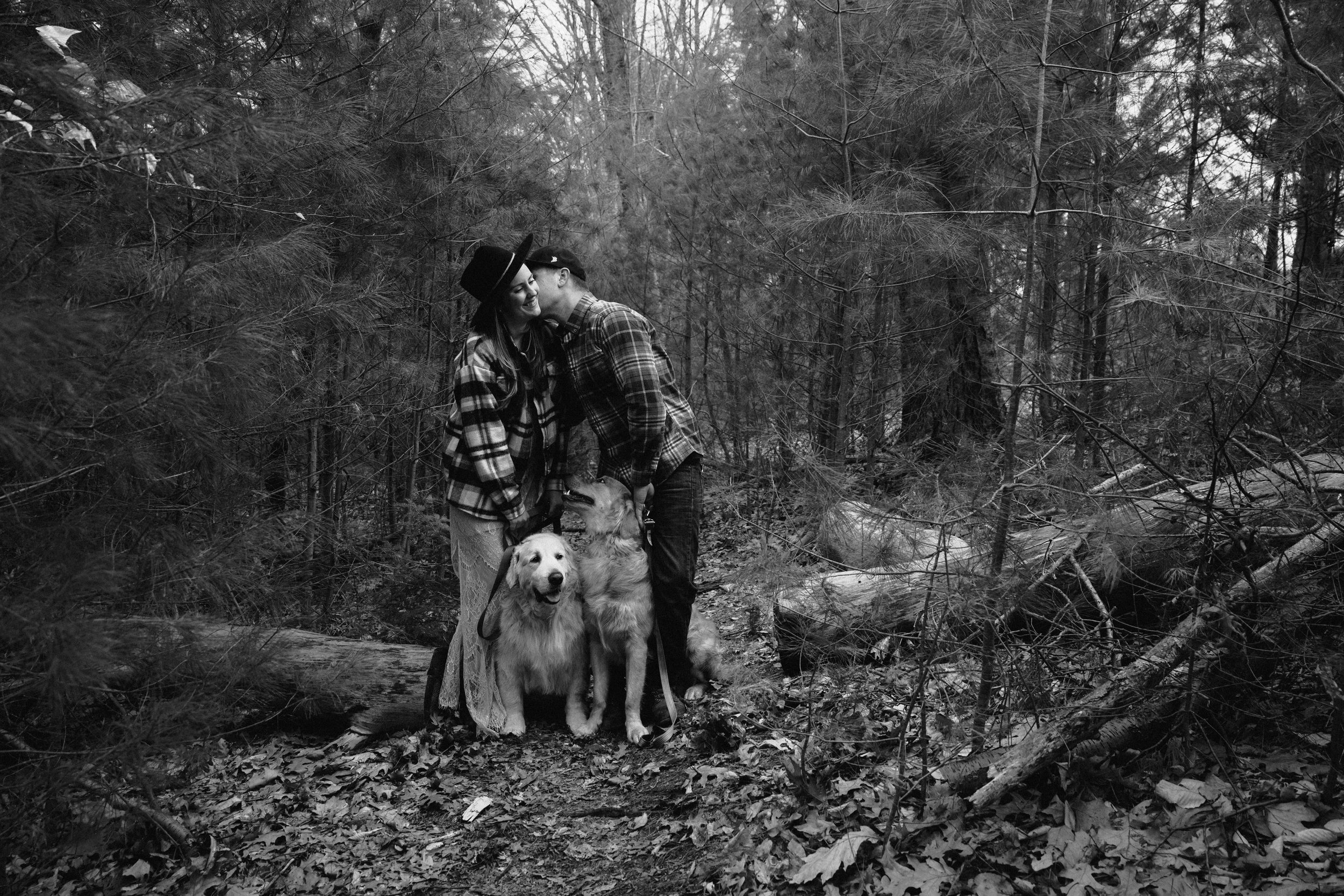 A black and white photo of a couple with two dogs in a dense forest, with one person kissing the other on the cheek.