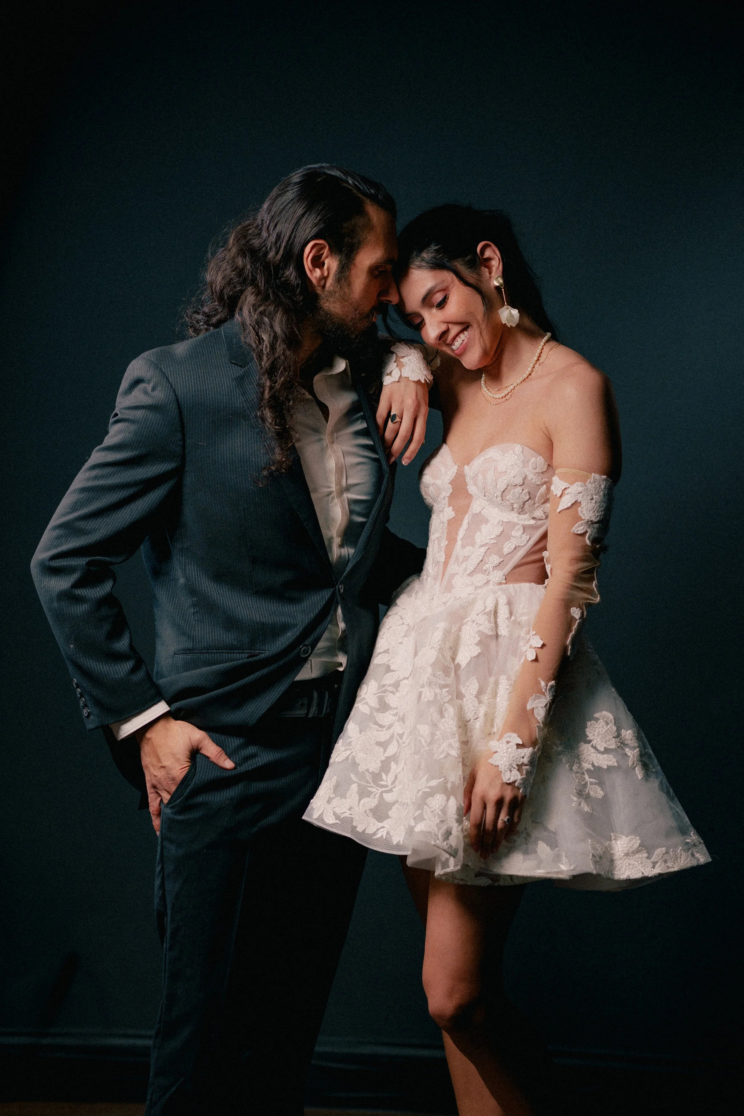 NJ Wedding Photography of A man and woman sharing an intimate moment, dressed in formal attire, with the woman wearing a strapless lace dress and the man wearing a dark suit, standing against a dark background.