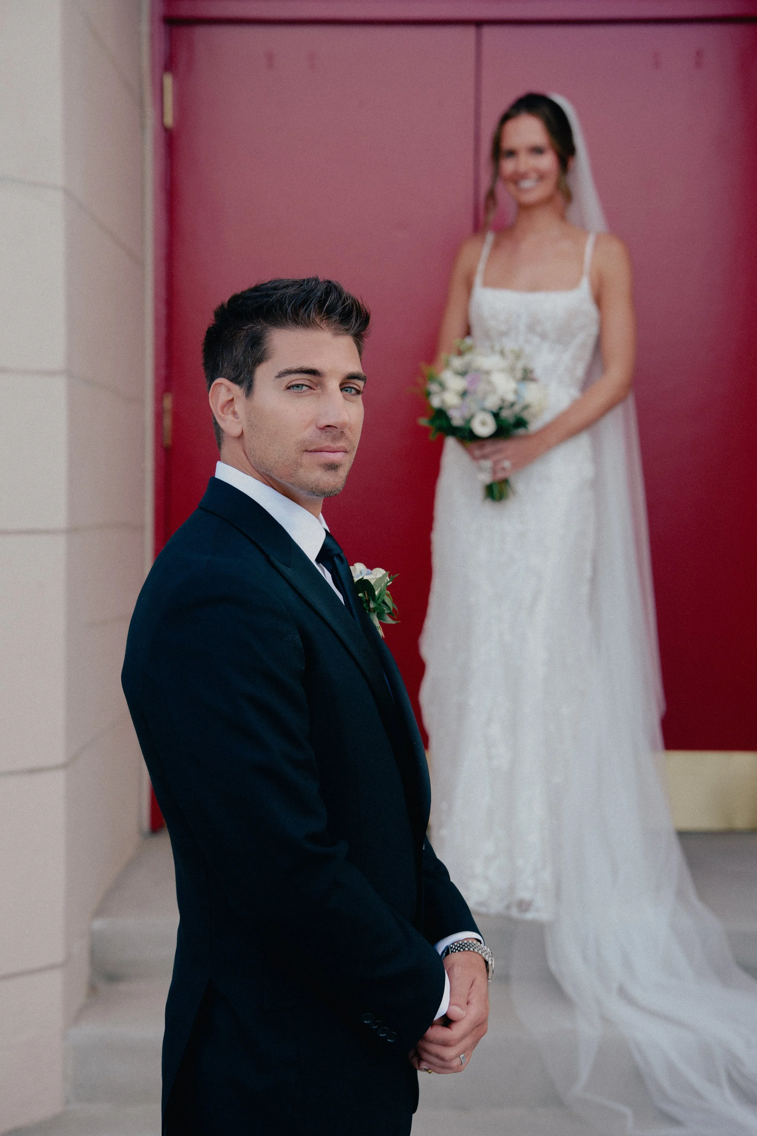 A groom in a dark suit with a boutonniere stands in front of a bride in a white wedding dress holding a bouquet, outside near a pink door at Wave resort - NJ Wedding photographers- Timeline Visuals
