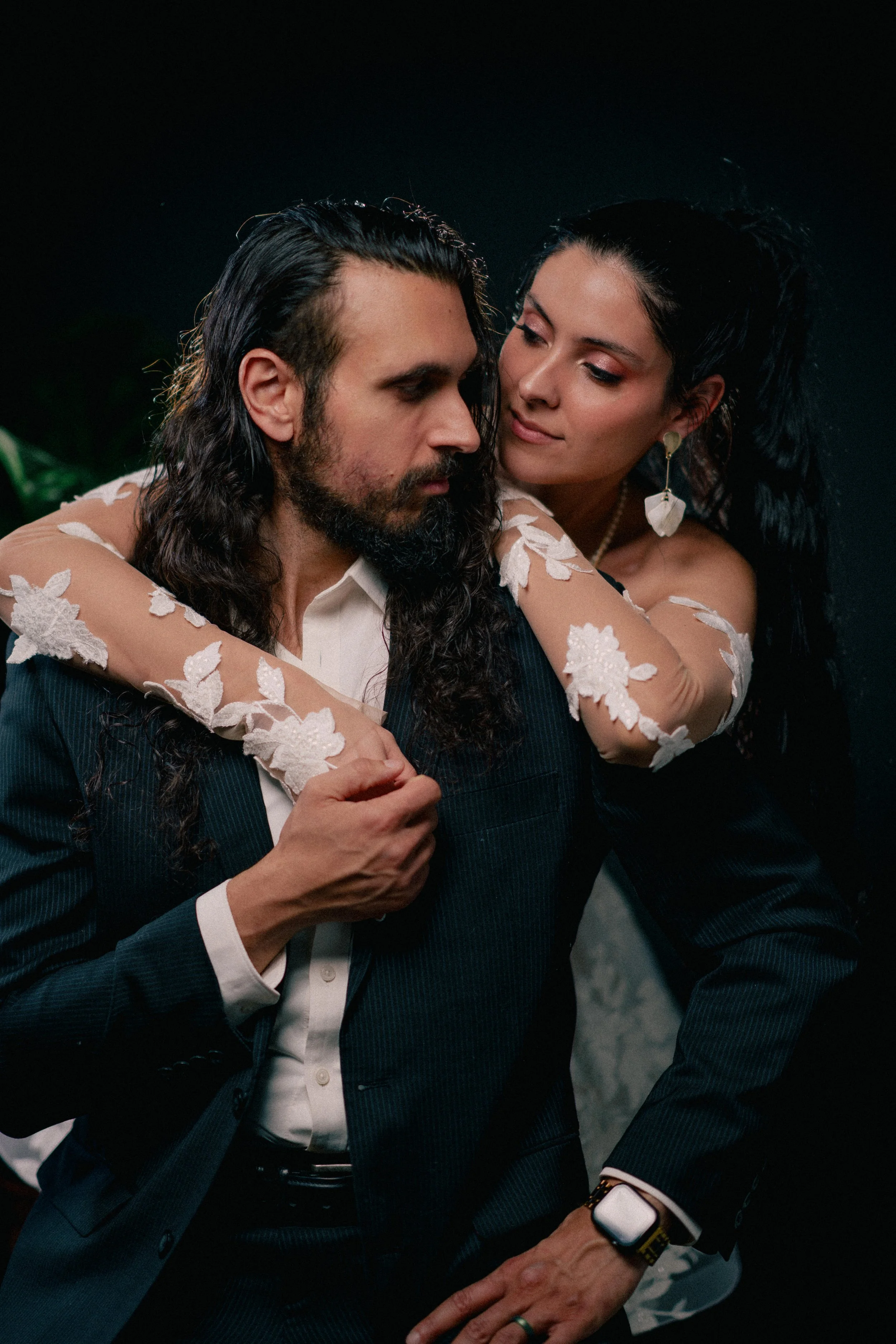 NJ Wedding Photography of A man in a suit with long dark hair and beard is being embraced by a woman with long dark hair wearing a dress with lace floral embroidery. The woman is standing behind him with her arms around his shoulders.