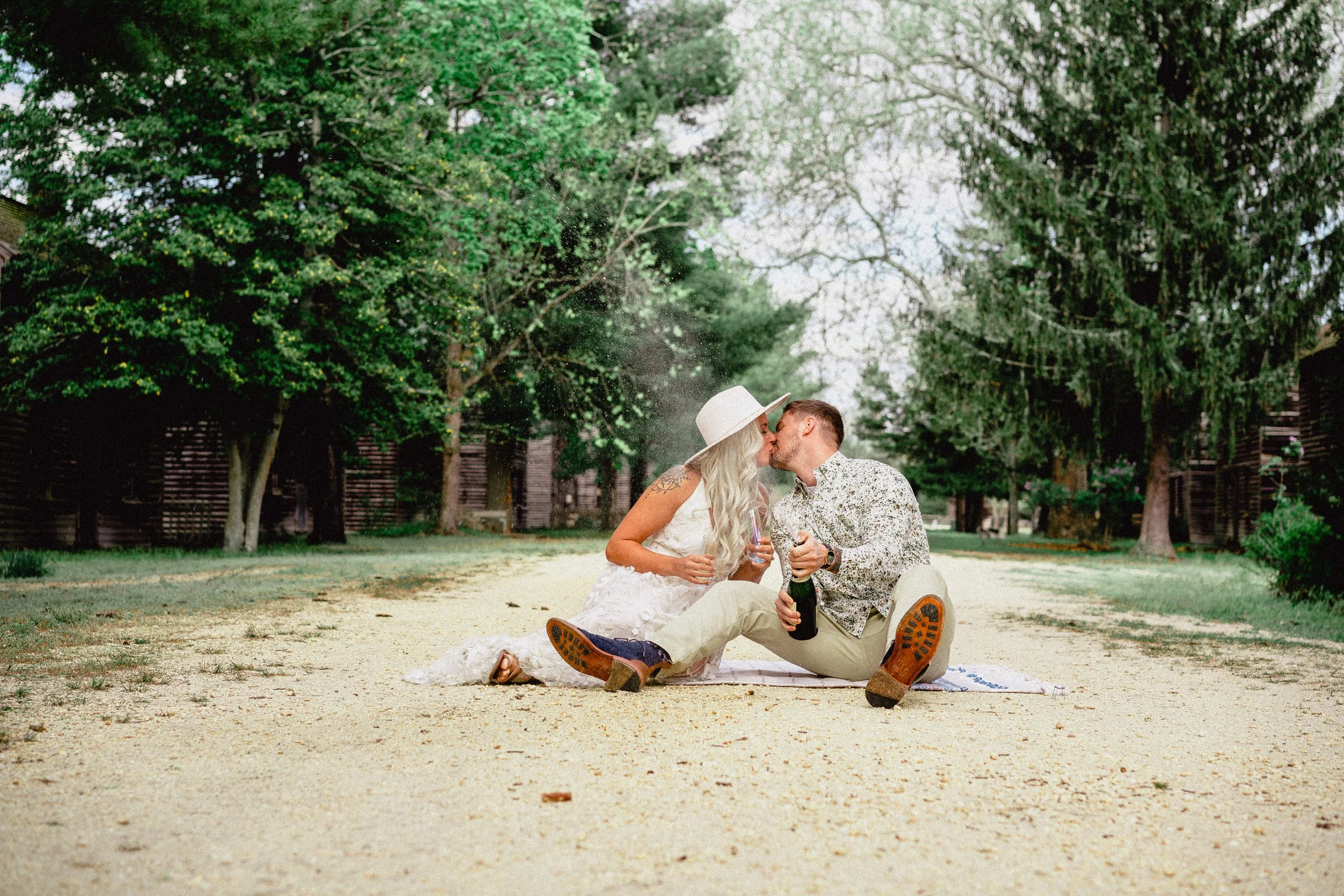 NJ Wedding Photography of A couple kissing while sitting on a blanket on a dirt path in a park, surrounded by green trees. The woman wears a white dress and hat, and the man wears a patterned shirt and beige pants. They are holding champagne bottles.