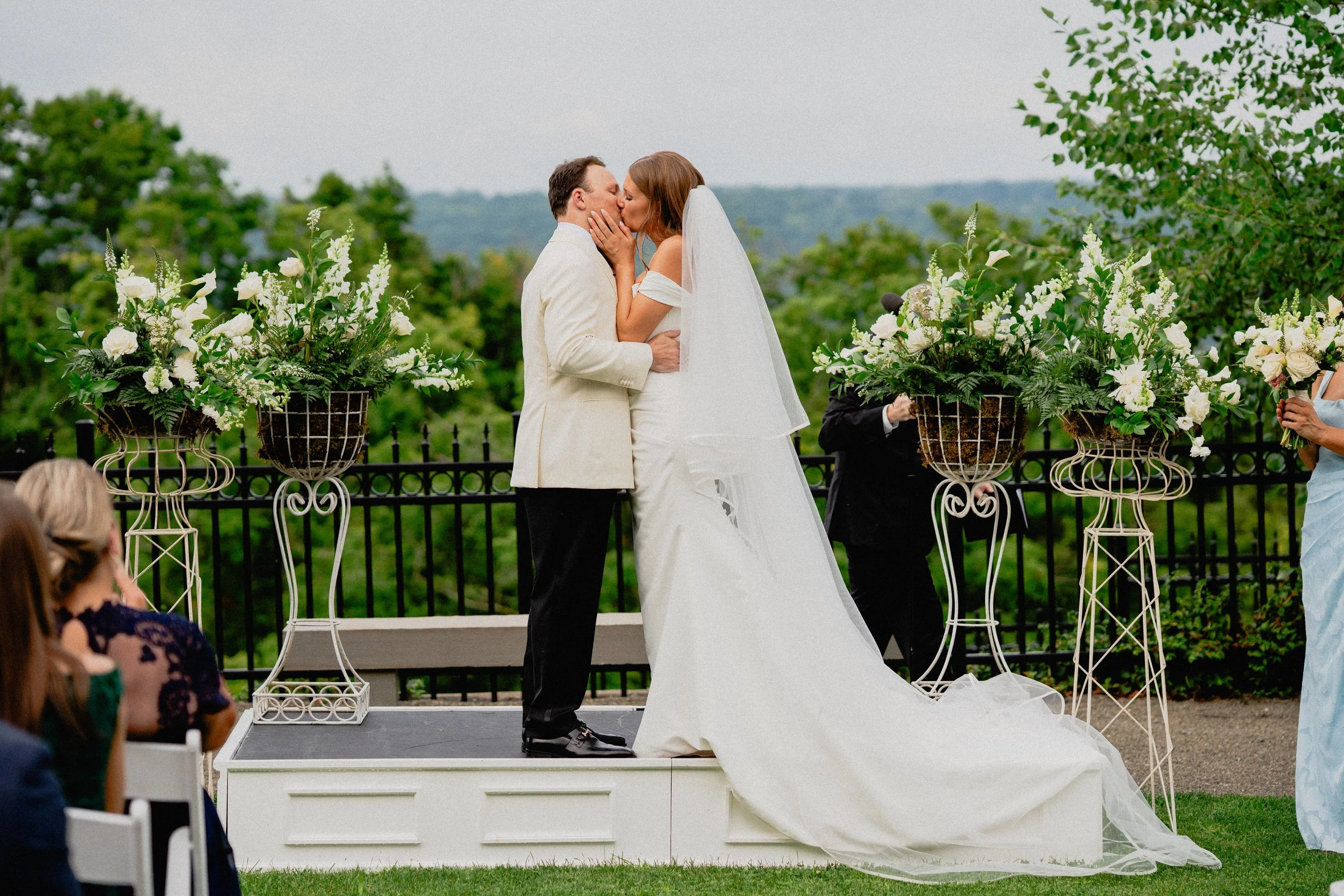 Luxury wedding photography of a bride and groom kissing beneath white flowers during their outdoor Upstate New York ceremony.