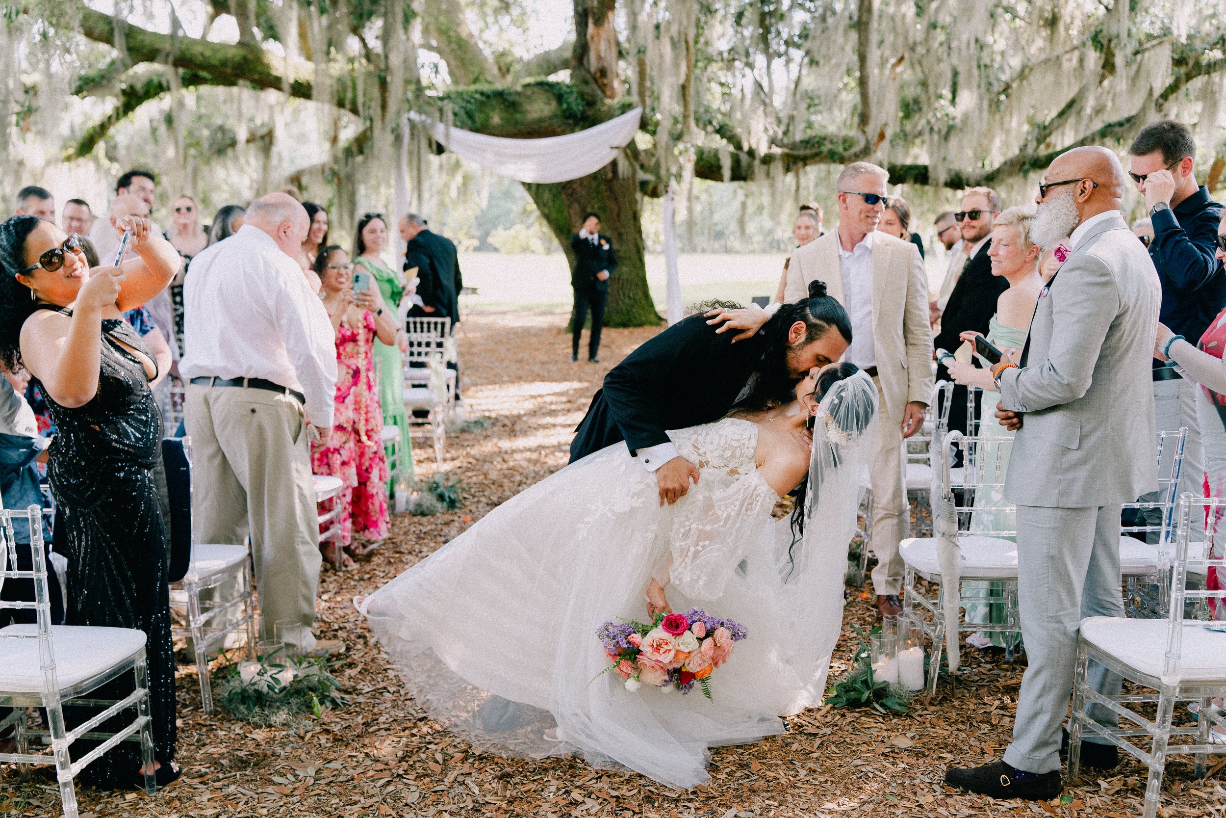 A wedding ceremony outdoors under a large moss-covered tree with a bride in a white gown and veil kissing a groom in a black suit. Guests are standing around, some taking photos and others smiling, with a mix of men and women dressed in formal attire