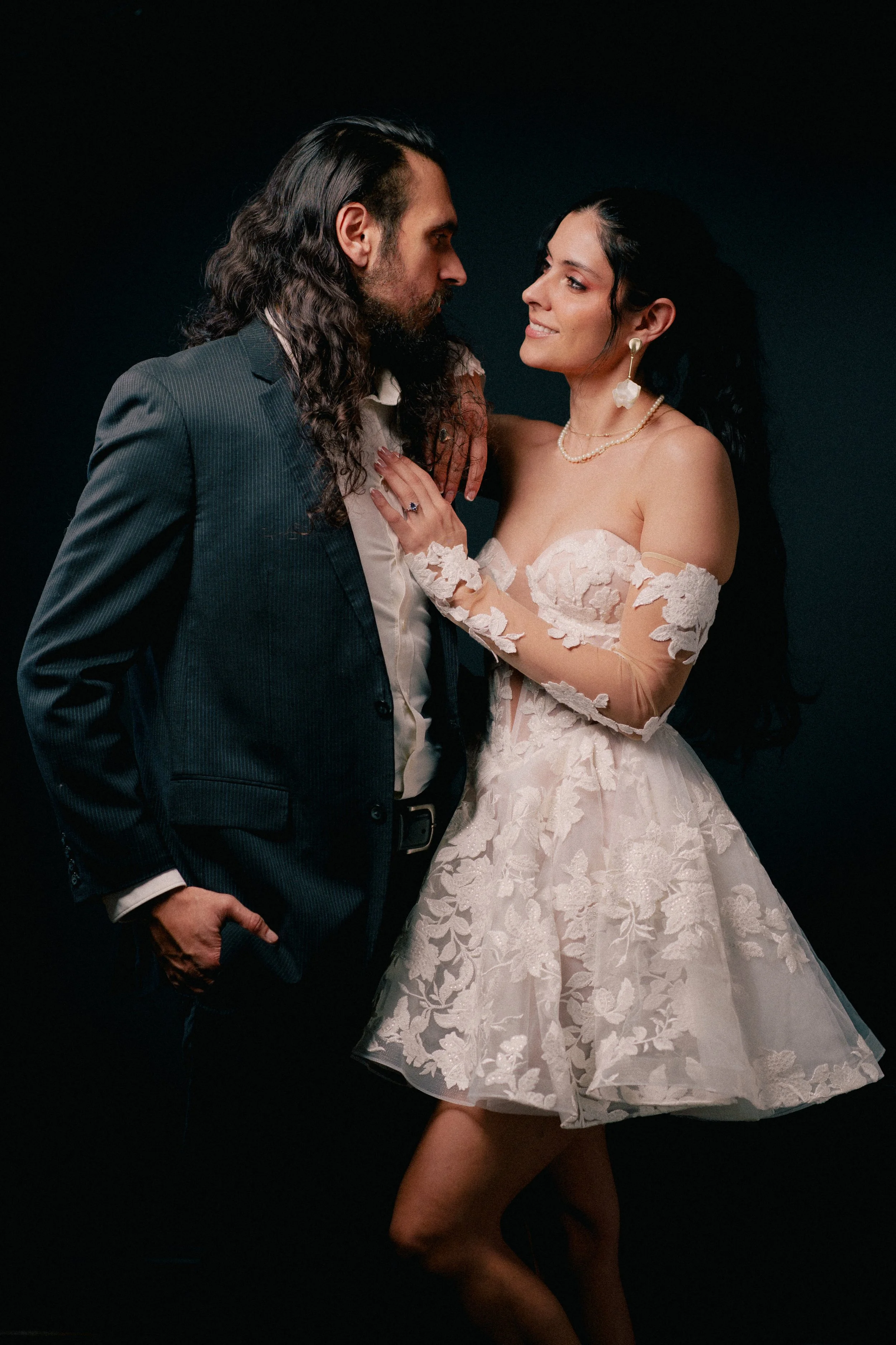 NJ Wedding Photography of A man and woman in formal attire looking at each other, with the woman wearing a lace off-shoulder dress and the man in a dark suit, against a dark background.