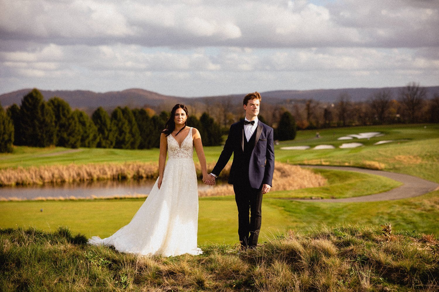 A bride and groom holding hands on a golf course with trees and hills in the background, under a partly cloudy sky.