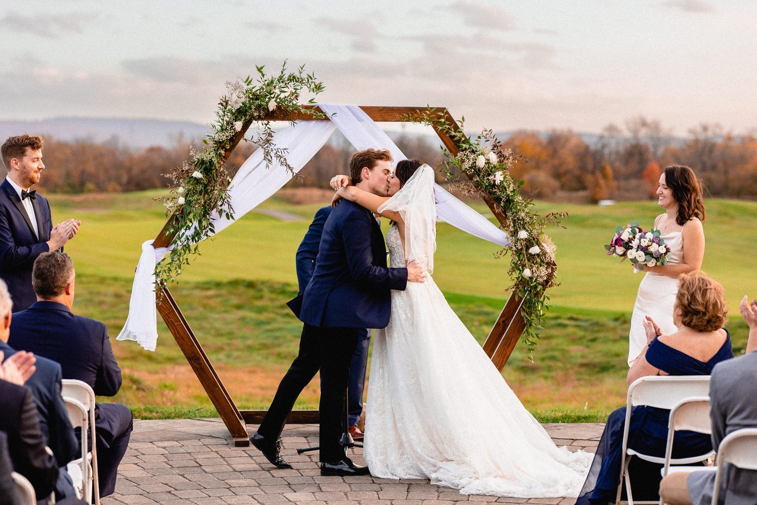 A wedding ceremony outdoors with a couple kissing at a decorated wooden arch, surrounded by seated guests, during sunset with an open field in the background.