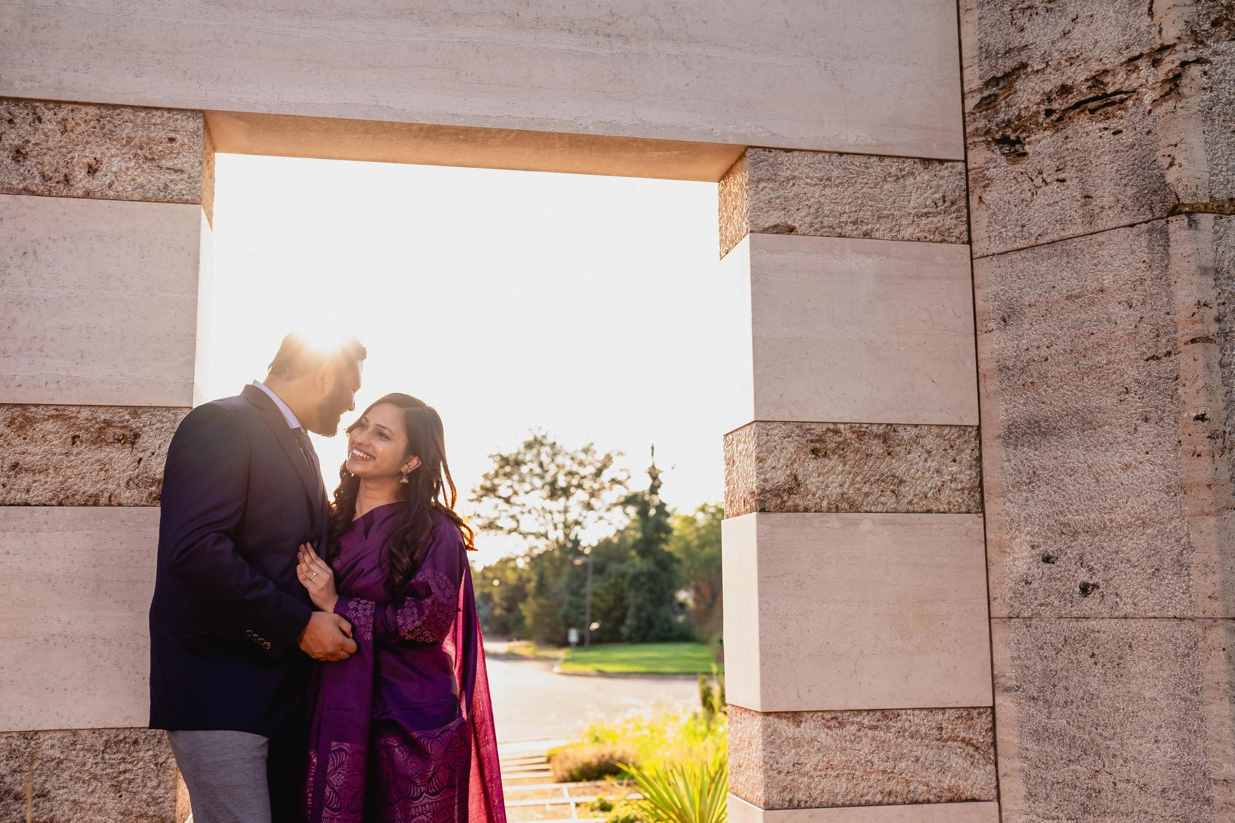 NJ Wedding Photography of A couple dressed in formal attire standing outdoors under a stone archway, smiling at each other, with sunlight shining behind them.