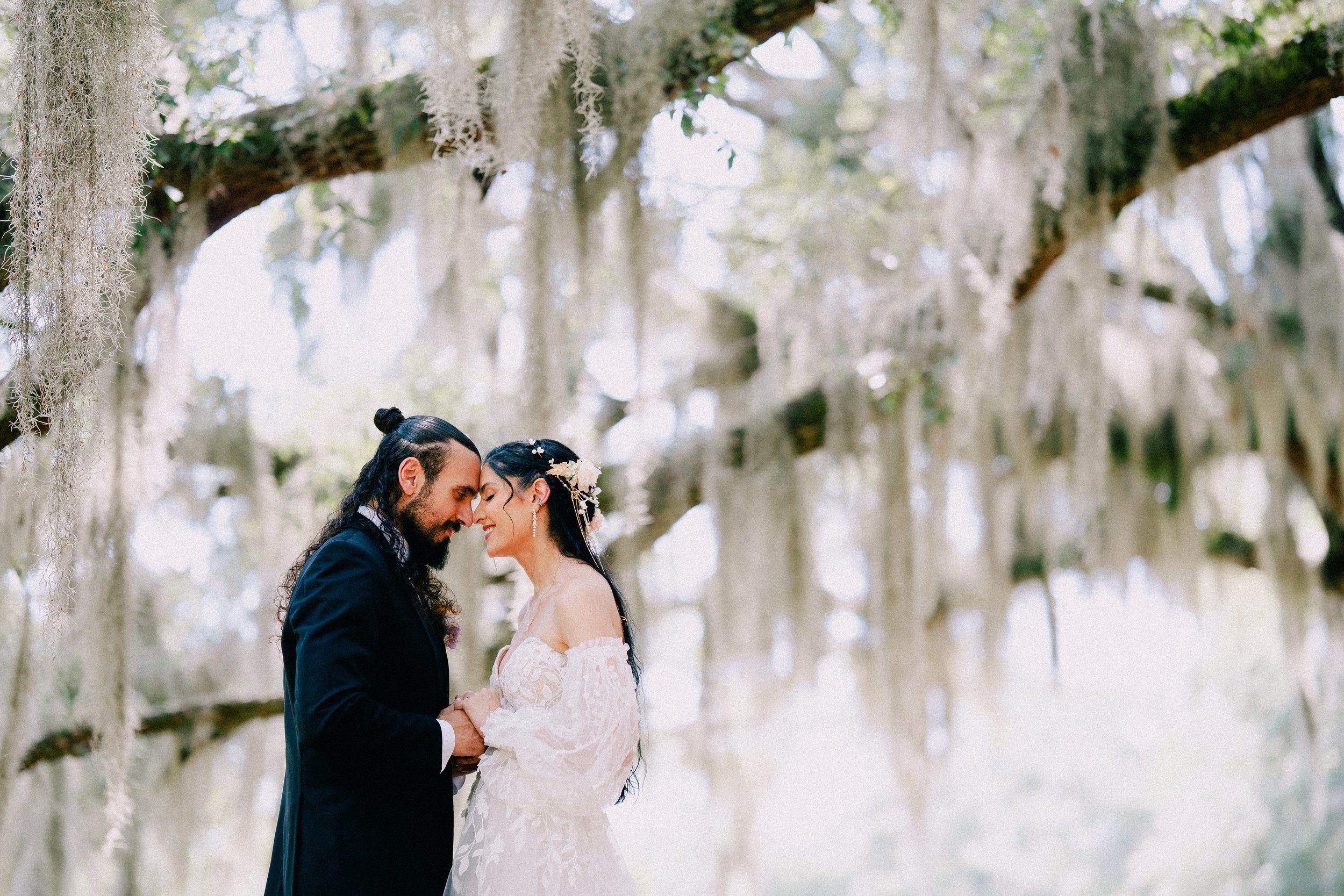 A couple in wedding attire sharing an intimate moment in a moss-covered forest, with their foreheads touching and eyes closed.