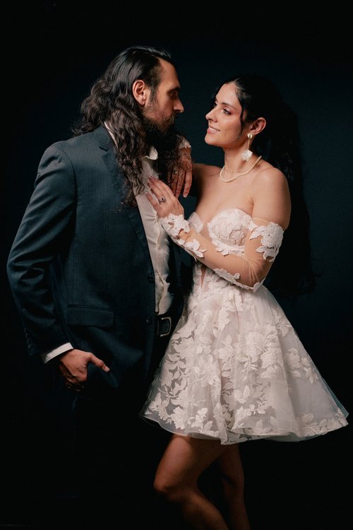 A man in a dark suit with long dark hair and a beard and a woman in a white floral dress with long dark hair and jewelry look at each other affectionately against a dark background.