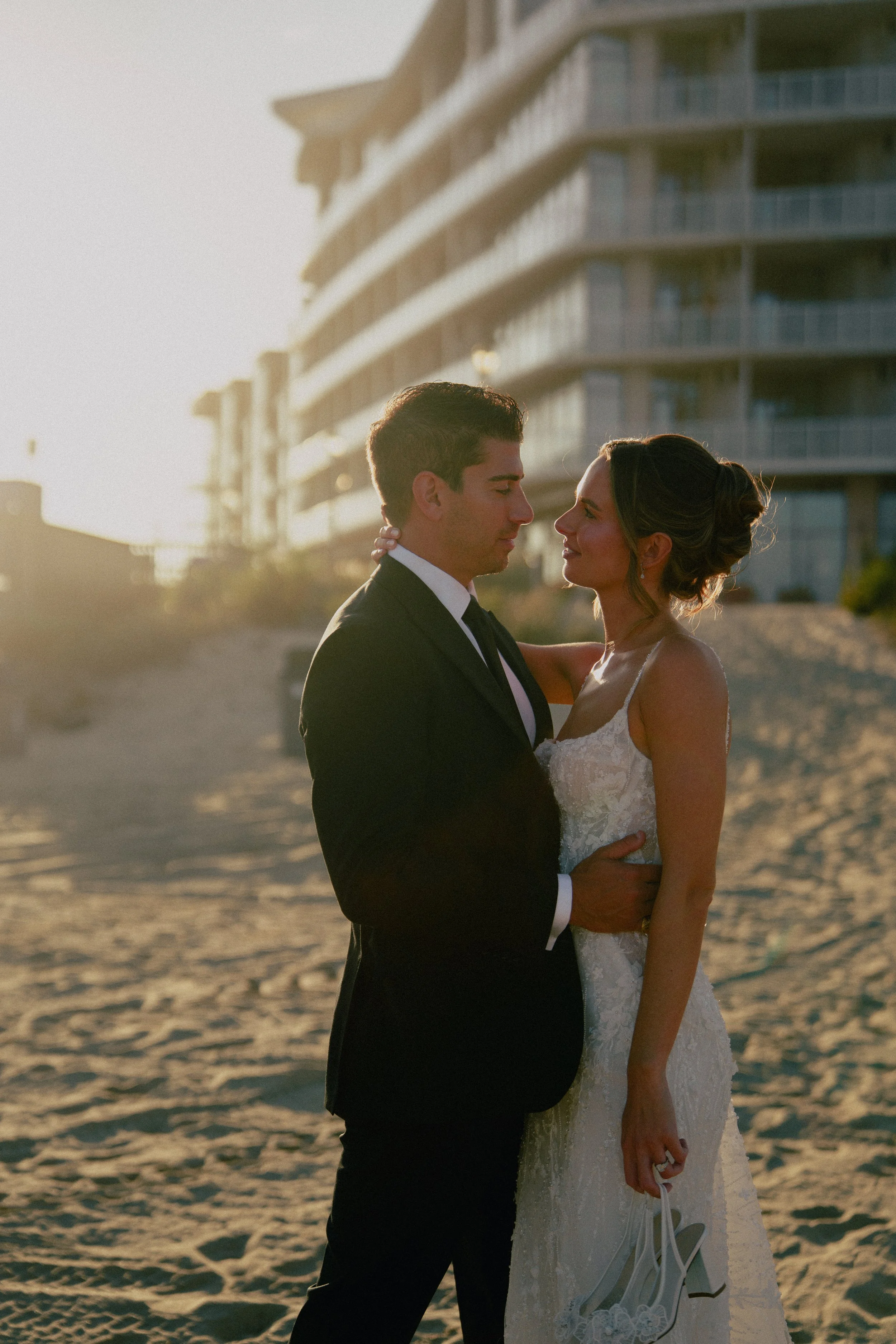 A NJ couple dressed in wedding attire on a beach at sunset, the man in a black suit and the woman in a white lace wedding dress, gazing into each other's eyes at wave resort - Wedding photographers- Timeline Visuals