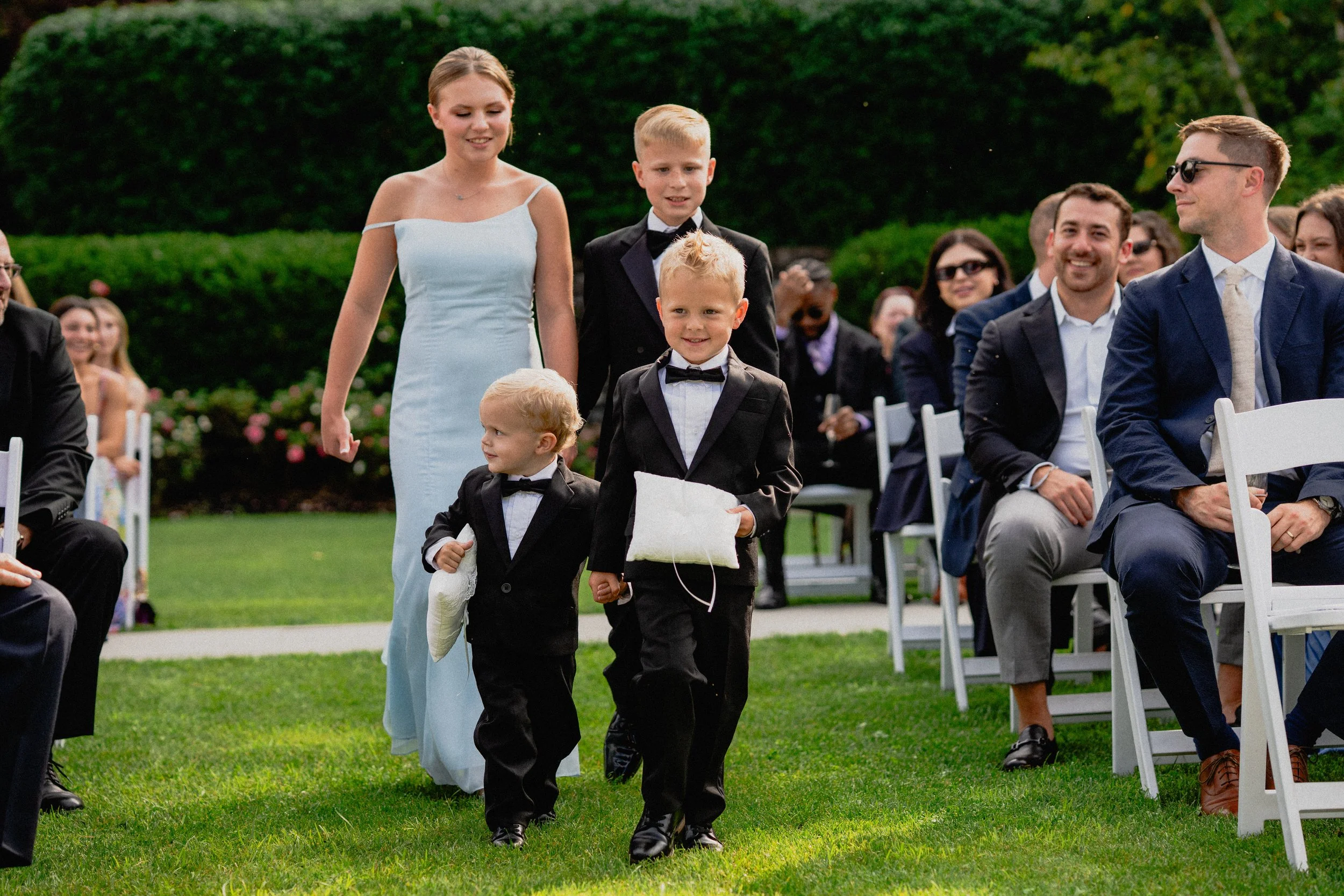 Documentary wedding photography of young ring bearers walking down the outdoor aisle with a woman in a floral dress.
