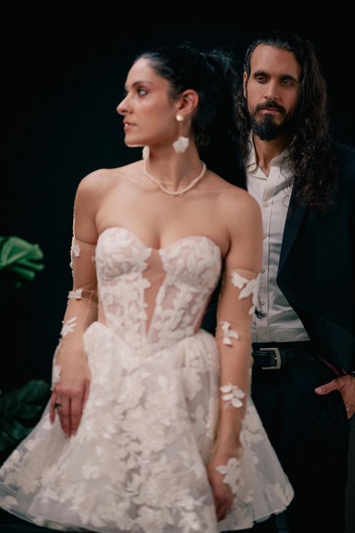 A woman in a wedding dress and a man in a suit stand together against a dark background. The woman wears a strapless, lace wedding gown with floral details, and the man has long hair, a beard, and is wearing a dark suit.