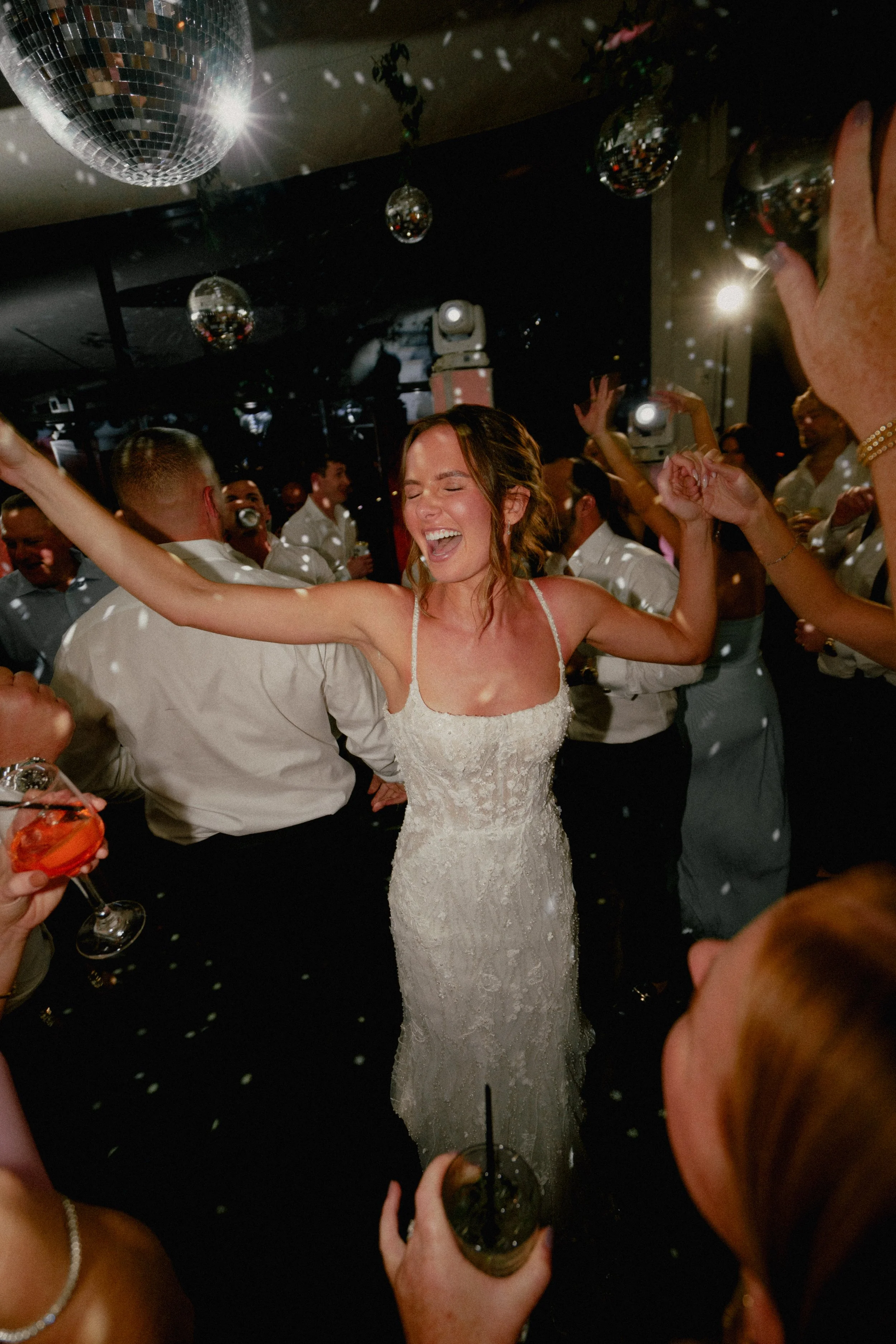 A nj bride in a white wedding dress dancing and smiling at a lively celebration with guests around her, disco balls overhead, and drinks in hand at Wave resort - NJ Wedding photographers- Timeline Visuals