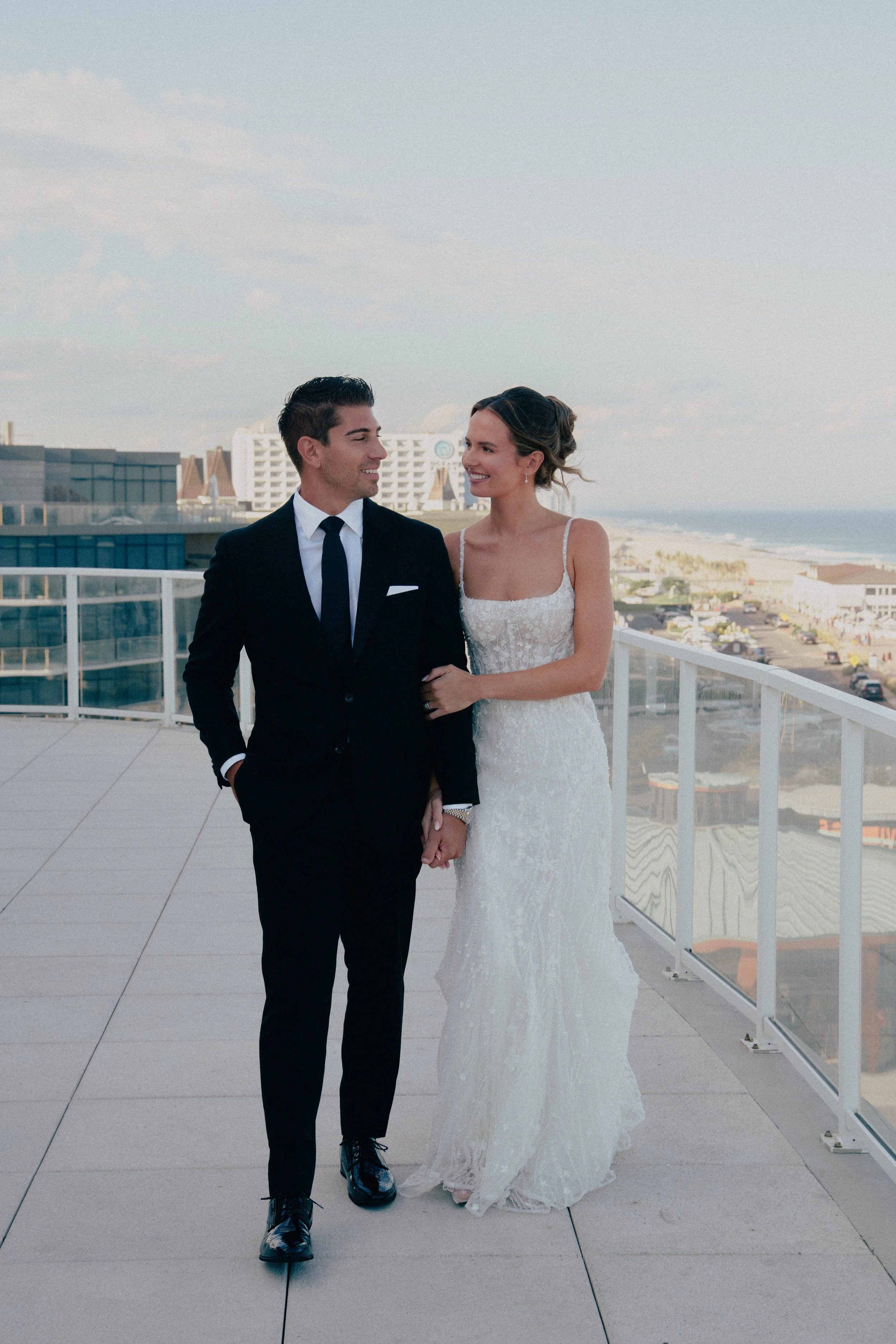A bride and groom walking hand in hand on a balcony with ocean view, the bride in a white lace gown and the groom in a black suit, smiling at each other at Wave resort - NJ Wedding photographers- Timeline Visuals