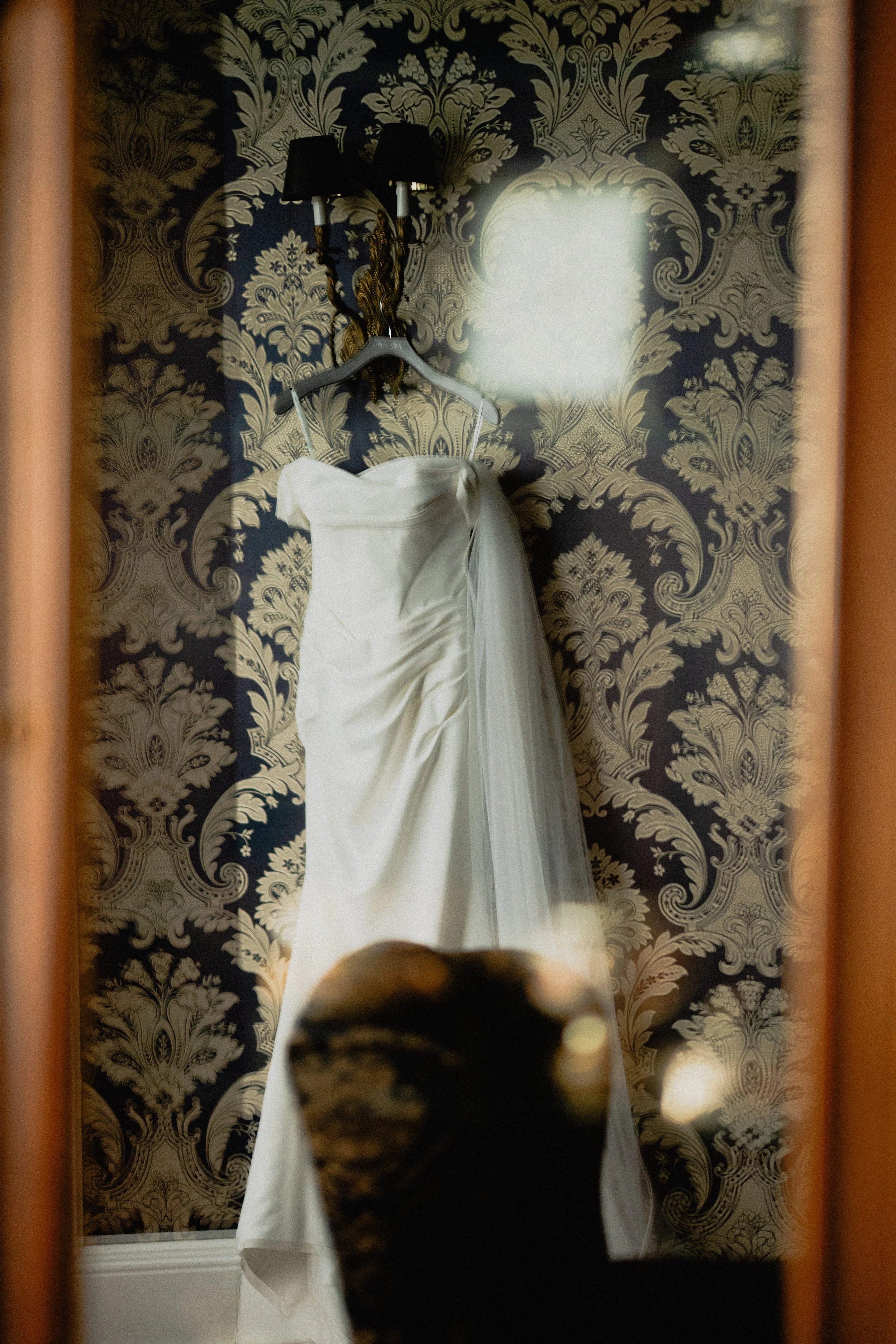 Editorial wedding photography detail of a bridal gown hanging against patterned wallpaper and reflected in a mirror.