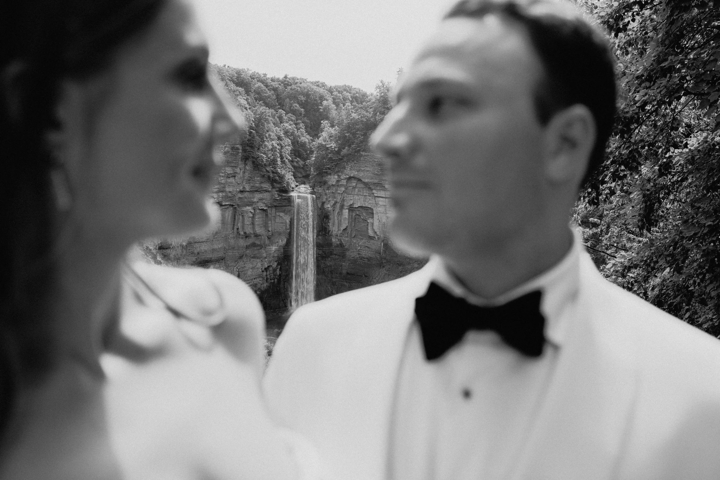 Black-and-white editorial wedding photography portrait of a couple at an overlook with a waterfall and green cliffs behind them.