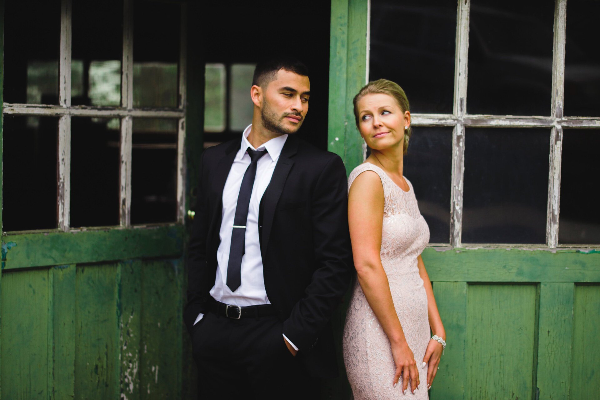 A man in a black suit and a woman in a light-colored lace dress standing close to each other against a rustic green and black barn or shed with windows. The man appears to be looking away while the woman looks at him with a slight smile.