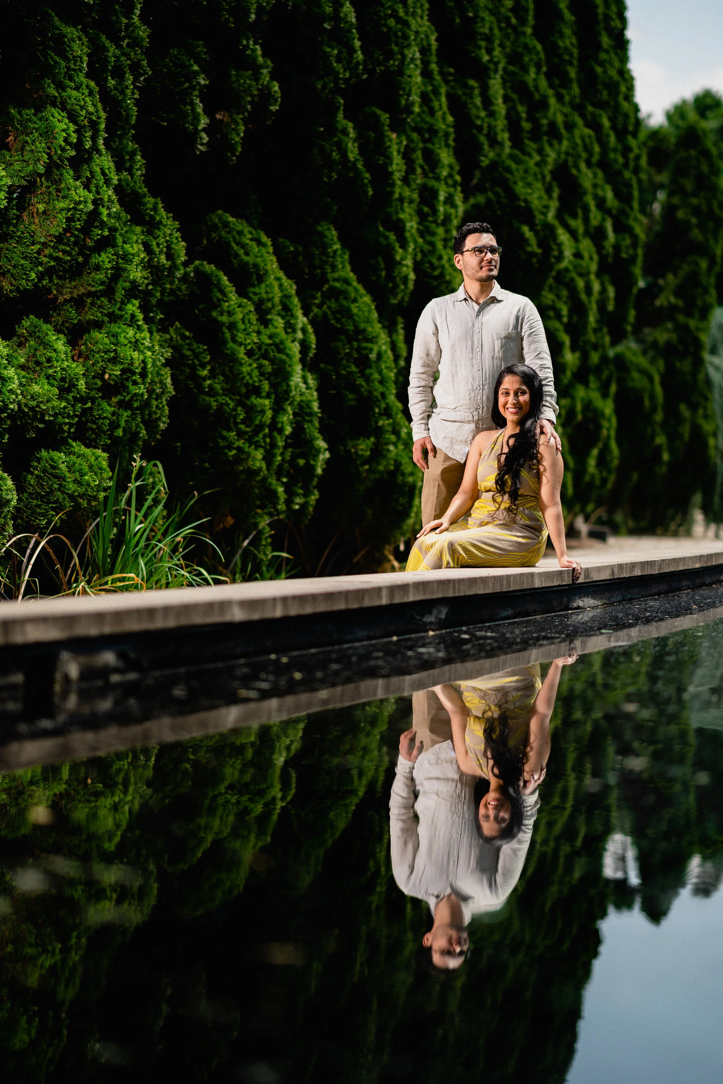 NJ Wedding Photography of A man and woman pose near a water feature with lush green bushes in the background, their reflection visible in the water.