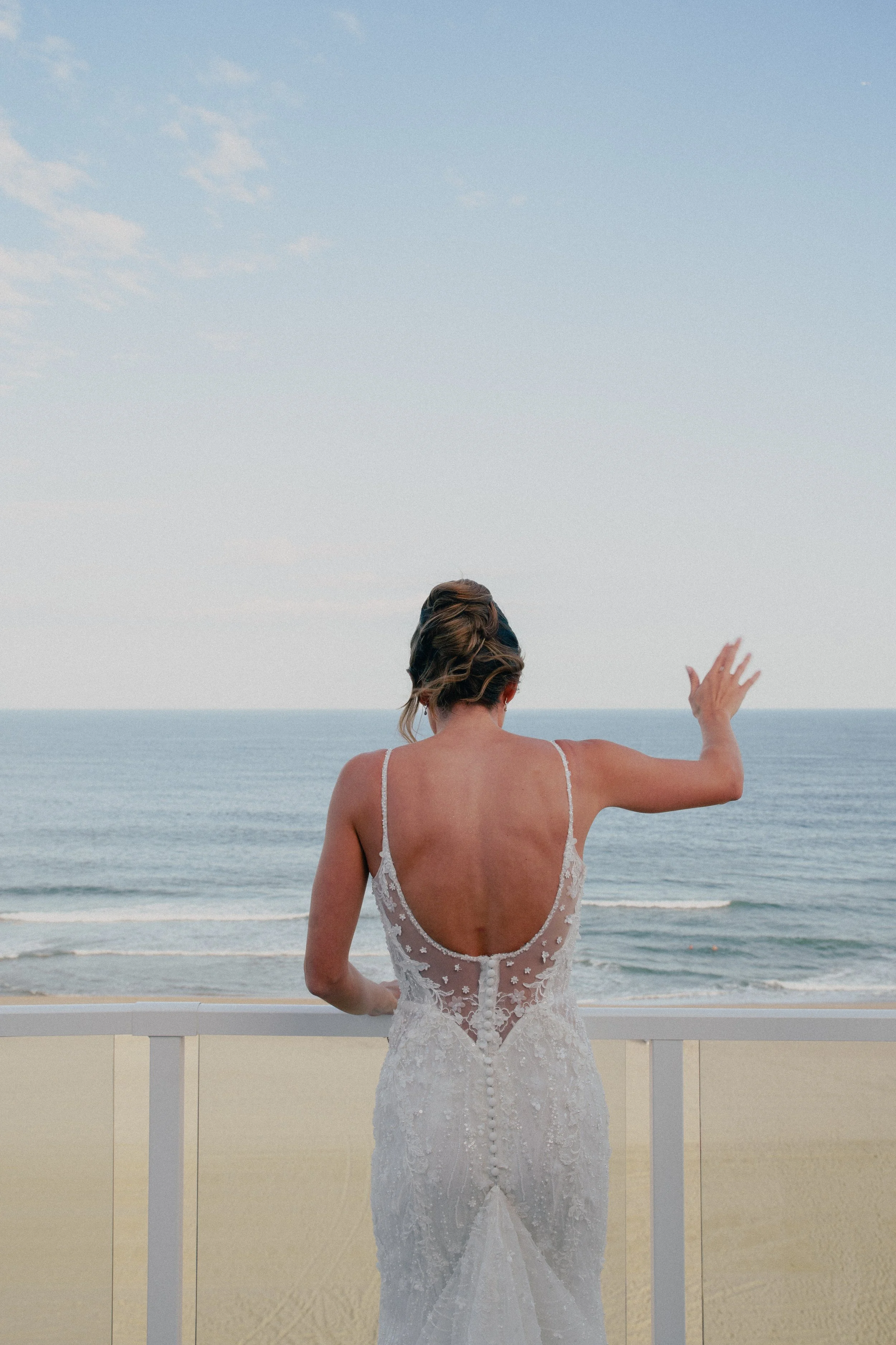 A NJ bride in her wedding dress with an open back stands on a balcony overlooking the beach and ocean, waving her hand at Wave resort - NJ Wedding photographers- Timeline Visuals