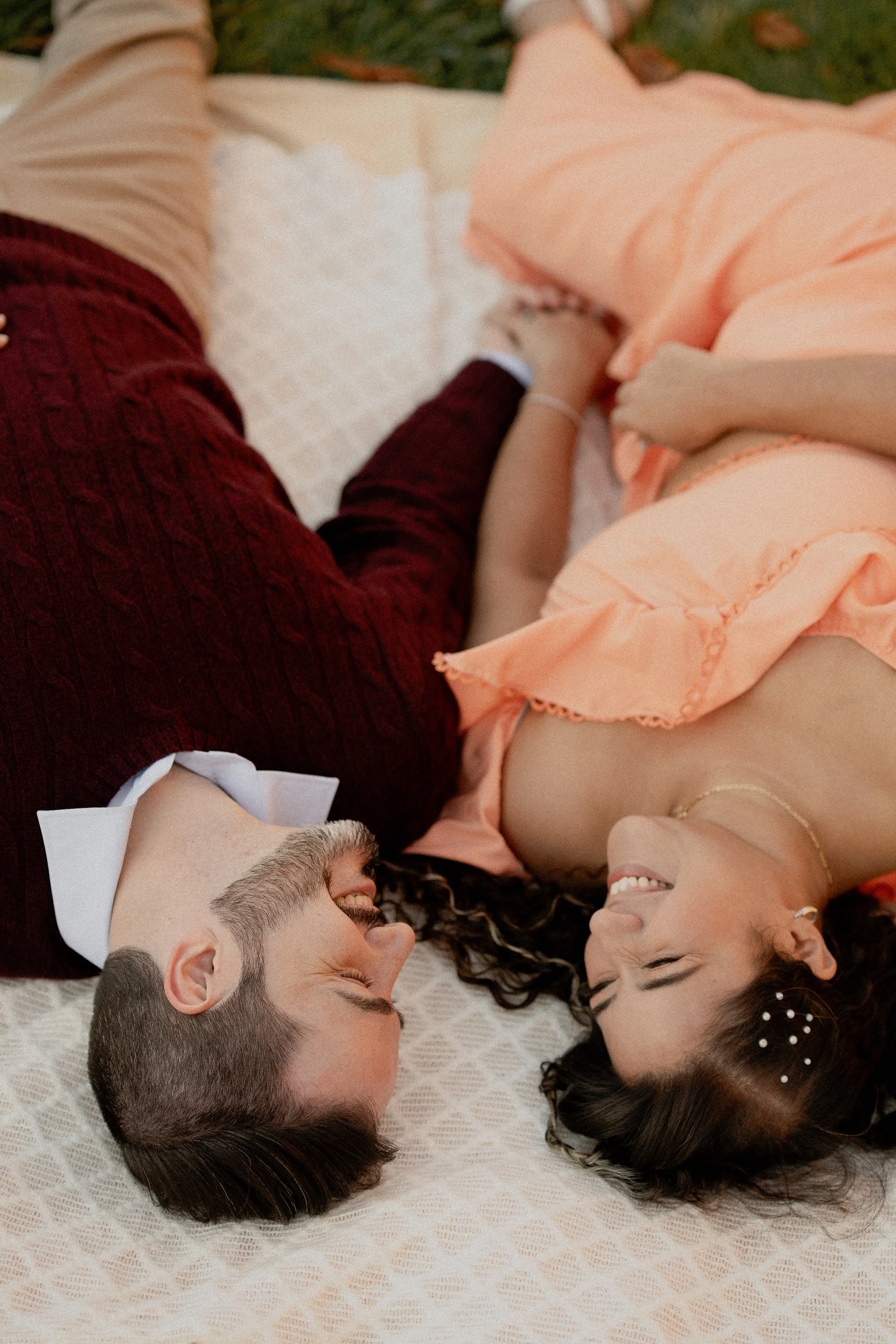 A man and woman lying on a bed, facing each other, smiling and holding hands. The man is wearing a tuxedo with a bowtie, and the woman is in a peach-colored dress with floral hair accessories, showing a joyous moment.