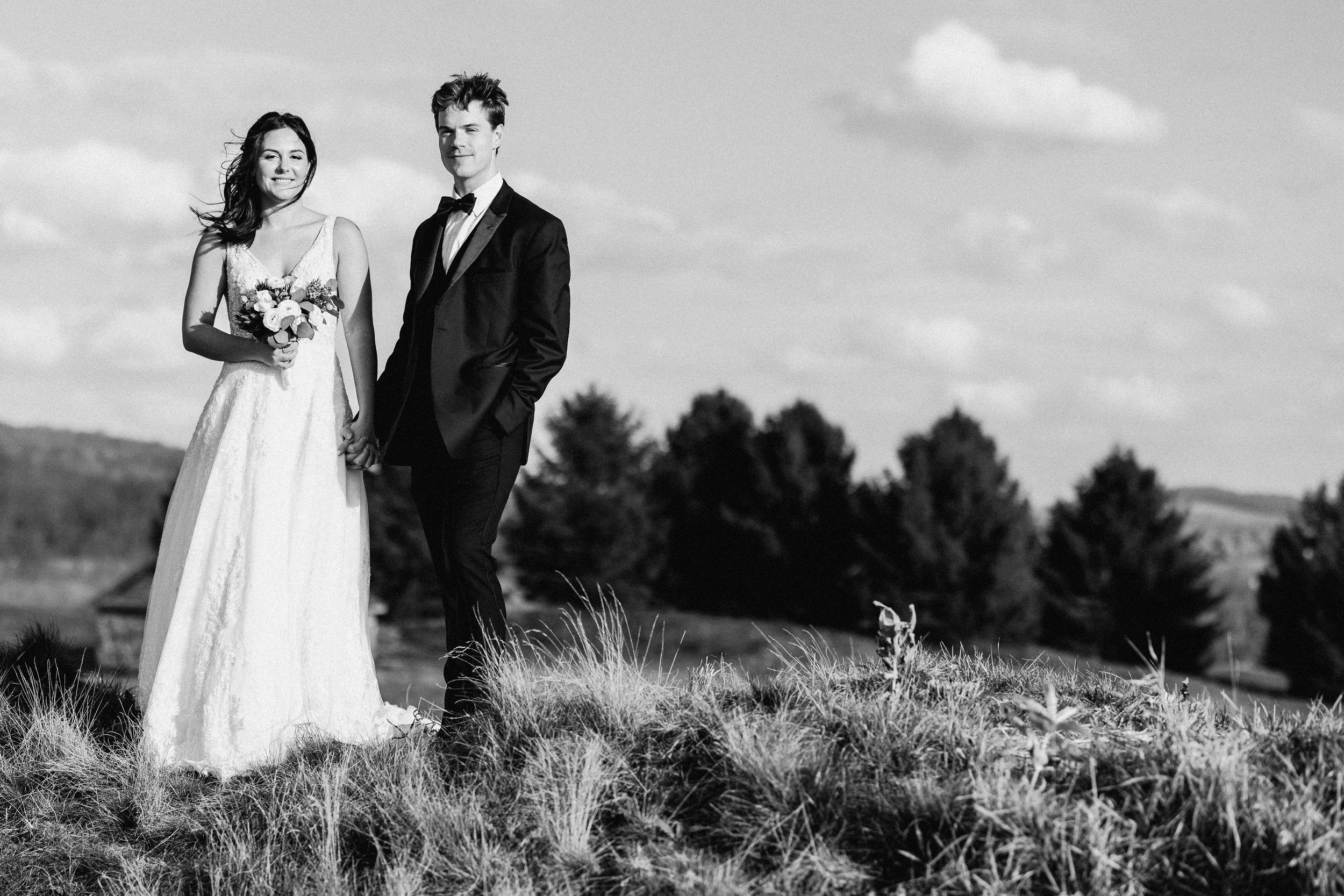 A bride and groom holding hands outdoors on a grassy hill, with trees and a partly cloudy sky in the background, captured in black and white.
