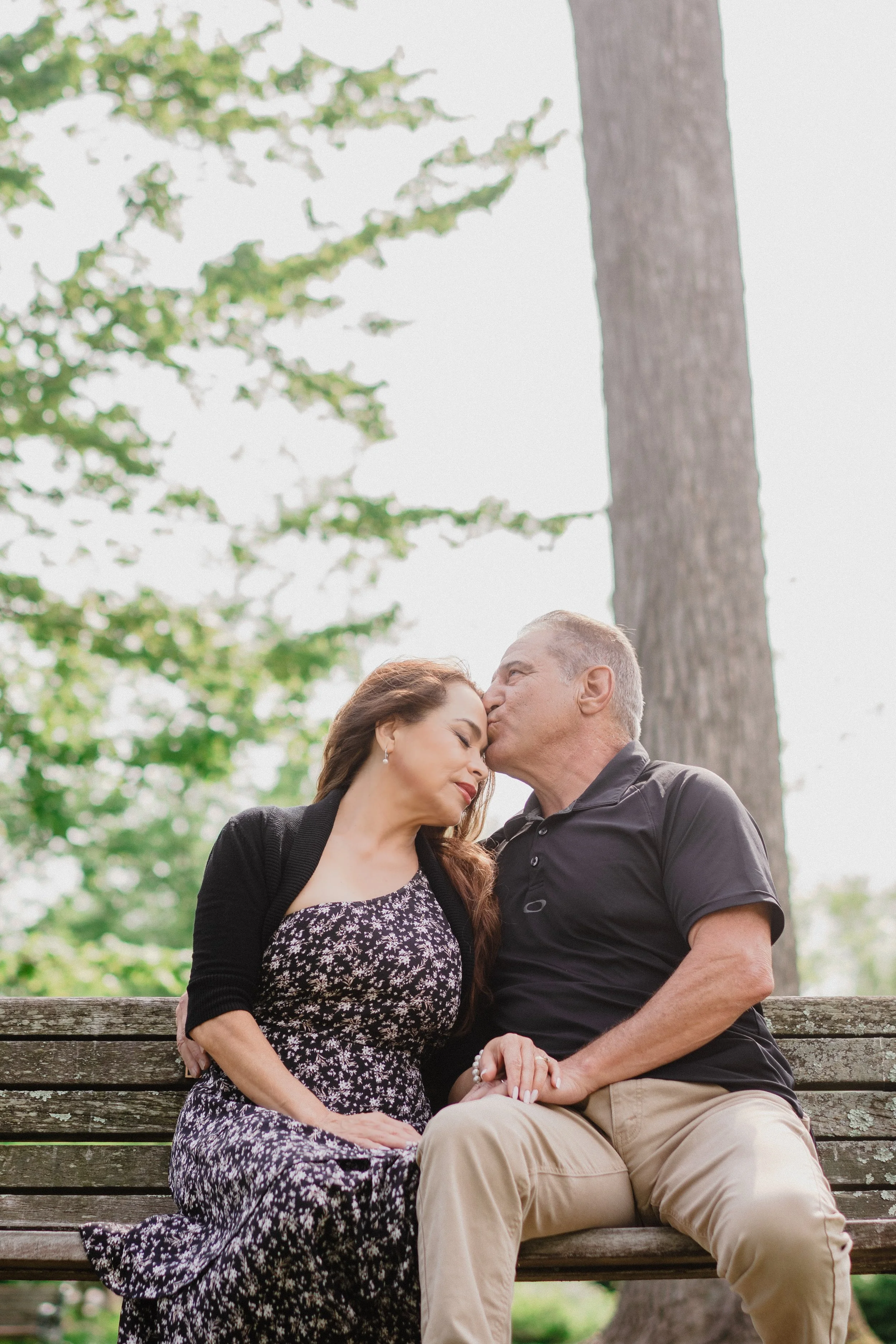 NJ Wedding Photography of A couple sitting on a park bench, the man kissing the woman's forehead, with trees in the background.
