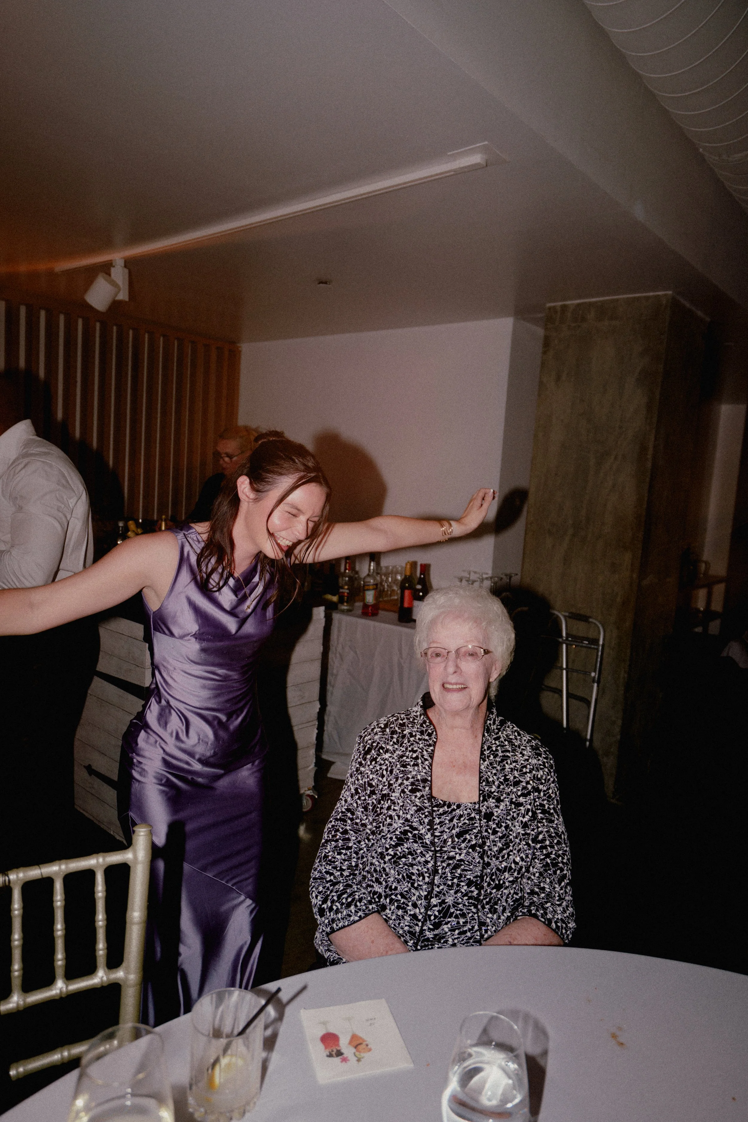 A young woman in a purple satin dress joyfully dancing with an elderly woman in a black and white patterned top, seated at a table during a celebration or party at Wave resort - NJ Wedding photographers- Timeline Visuals