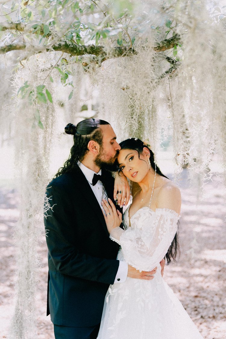 A bride and groom embrace under hanging Spanish moss and tree branches in an outdoor setting, with the groom kissing the bride's forehead.