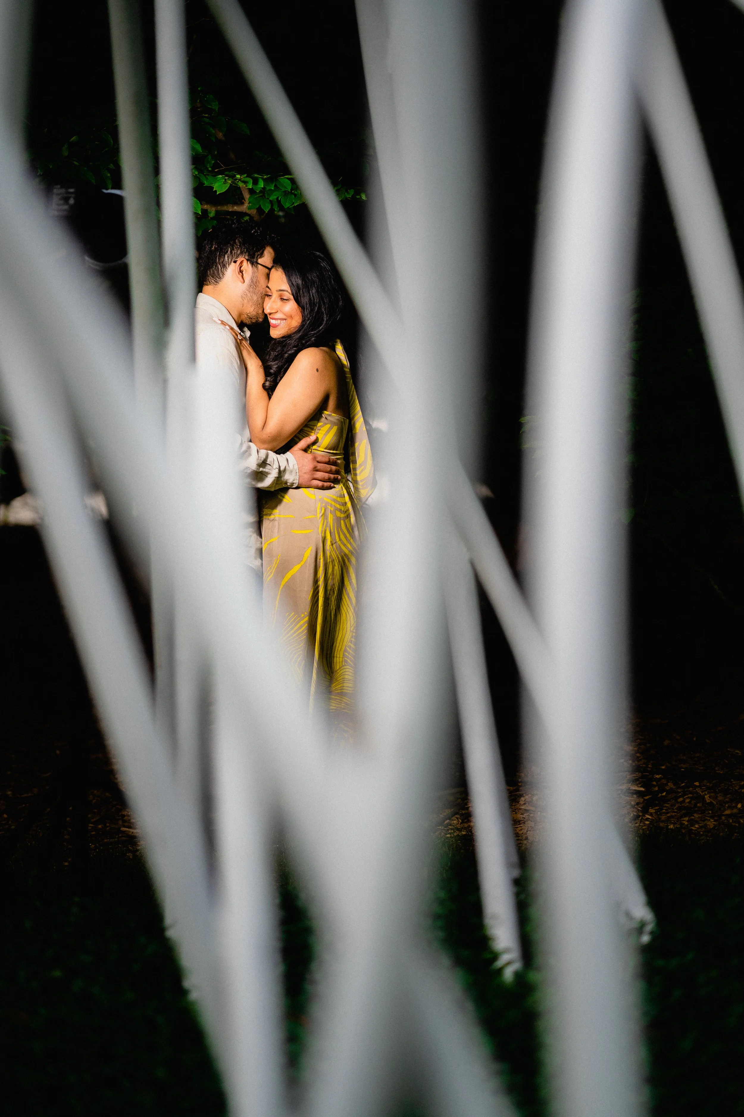 NJ Wedding Photography of A couple kissing outdoors at night, viewed through white metal bars or fence.