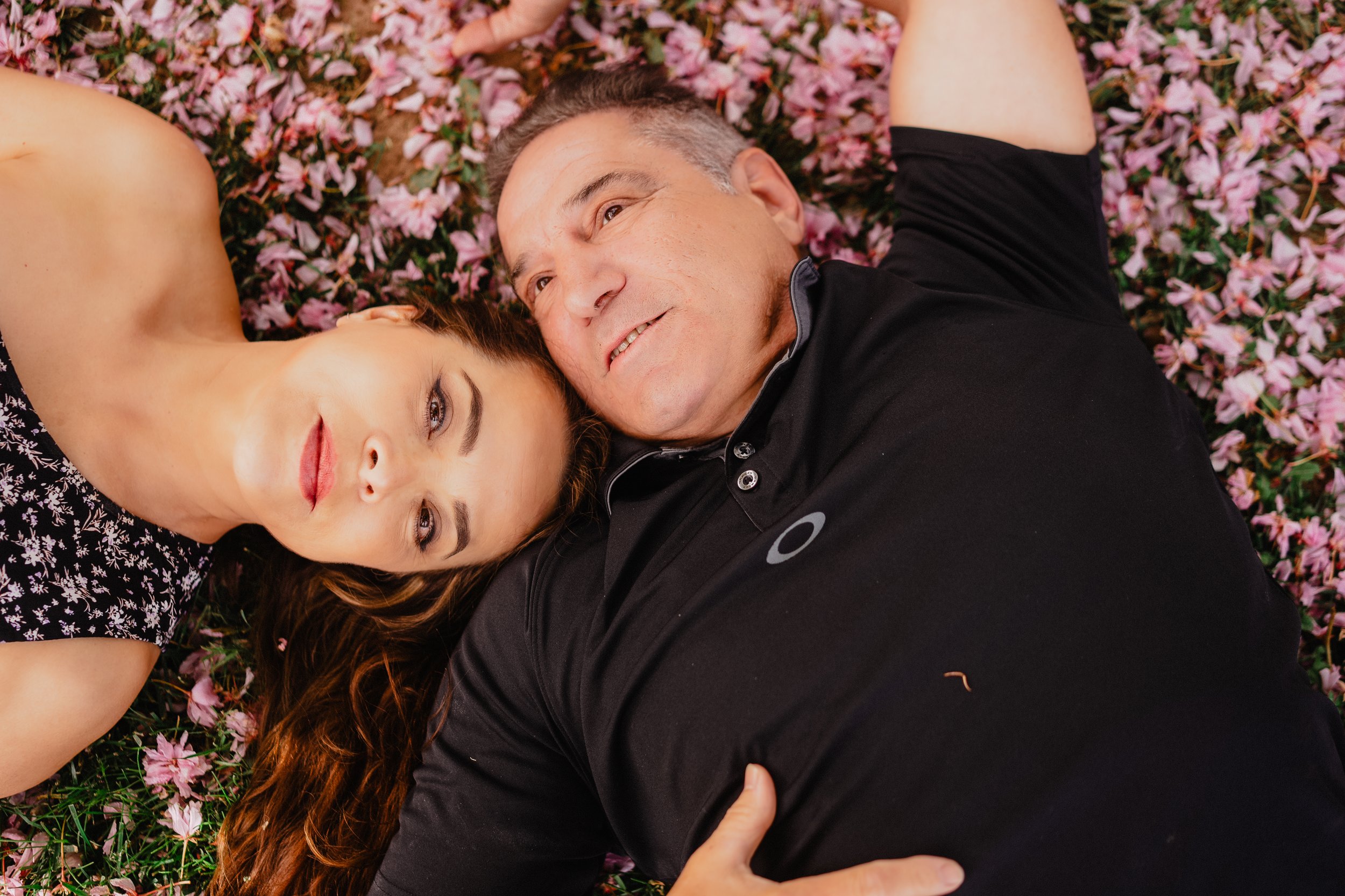 NJ Wedding Photography of A woman and an older man lying on a bed of pink flowers, looking at the camera, with the woman's head resting near the man's face.