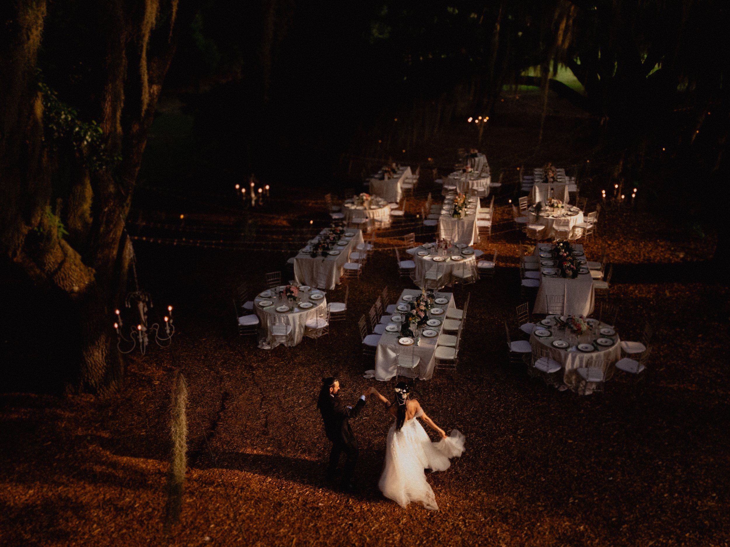 A bride and groom dancing outdoors at their wedding reception under a large, shadowy tree, with round and rectangular tables set with elegant white tablecloths, floral centerpieces, and plates, amid candlelight at night.